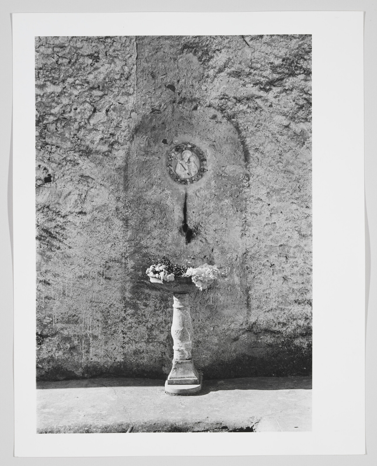 A stone pedestal with a shallow bowl of flowers stands against a weathered wall with a circular relief.