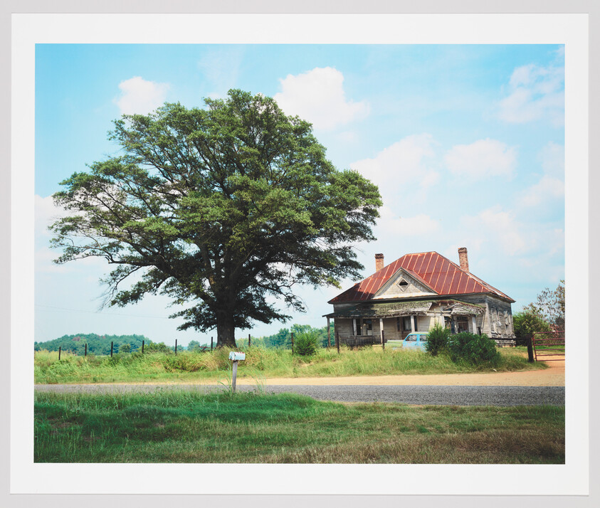 A large tree stands to the left of an old, dilapidated house with a rusted red roof, situated beside a country road with a mailbox in the foreground. The sky is partly cloudy, suggesting a warm, serene day.