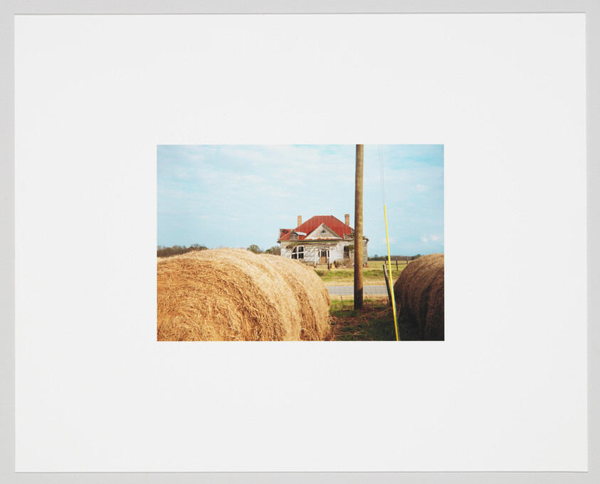 A photograph of a rural landscape featuring large round hay bales in the foreground and a classic red-roofed farmhouse in the background, with a clear blue sky above and a utility pole to the right. The image is bordered by a wide white frame, suggesting it is a printed photo or a picture within a mat board.