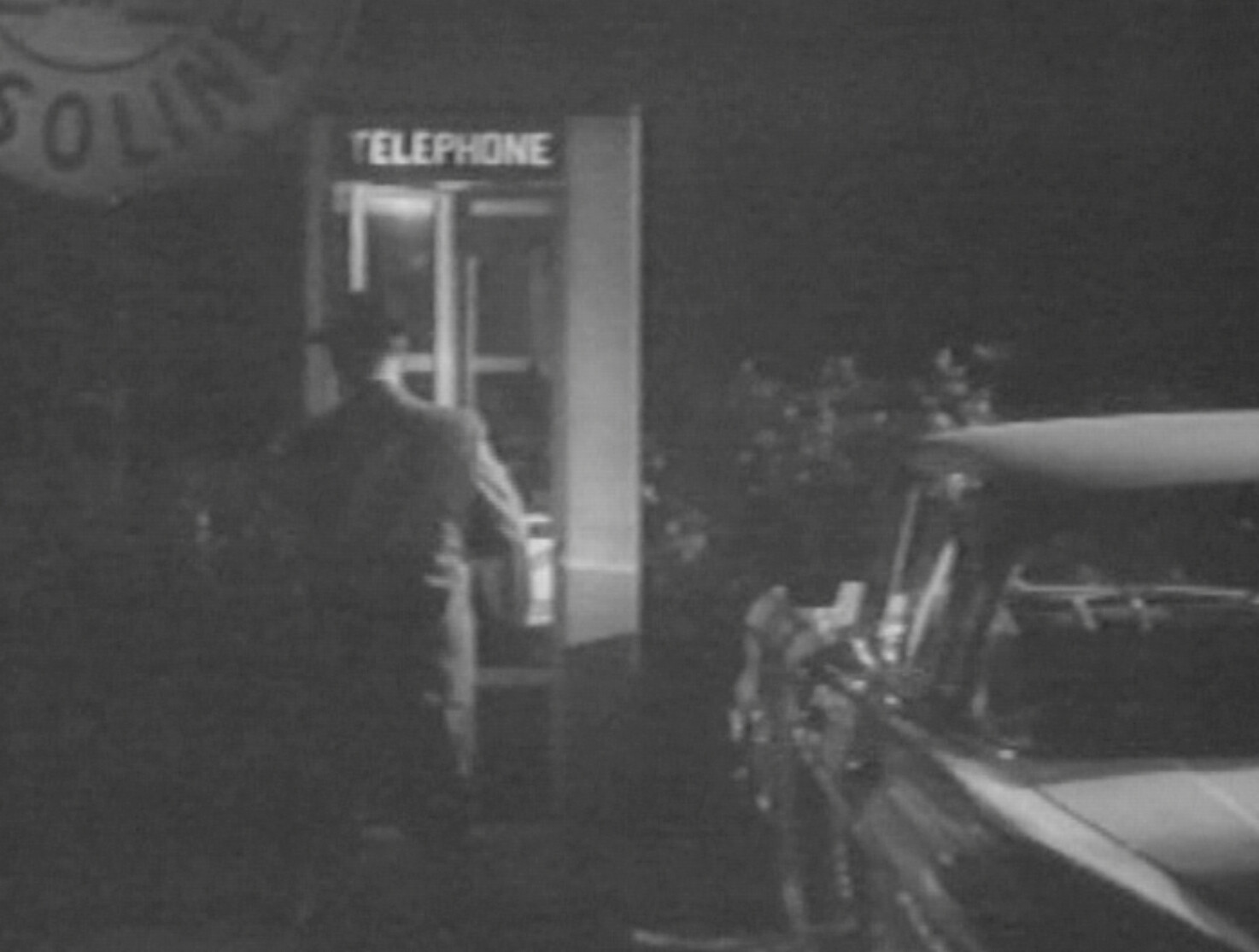 Man enters a lit telephone booth at night next to a parked car.