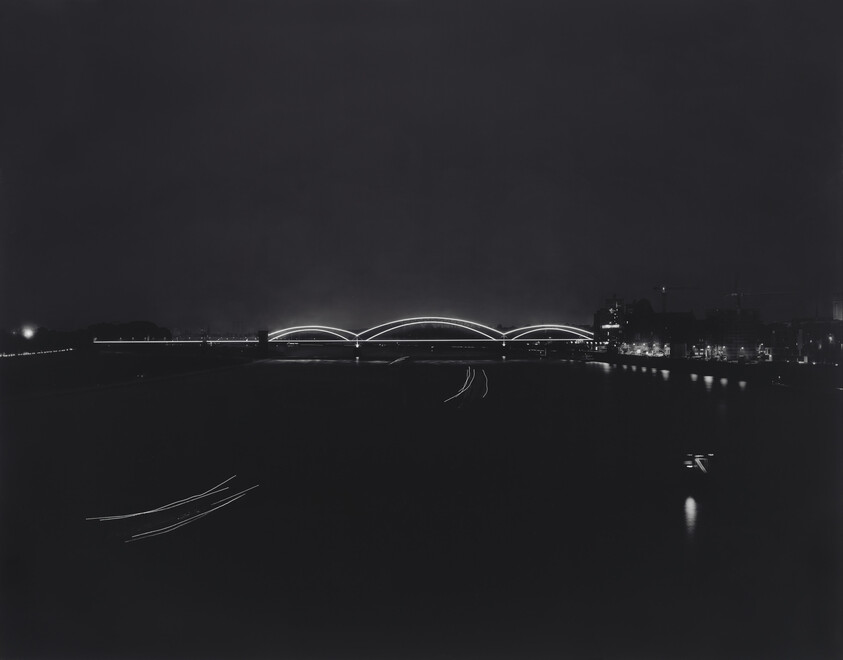 A lit arched bridge glows over a river at night with boat light trails and distant city lights.