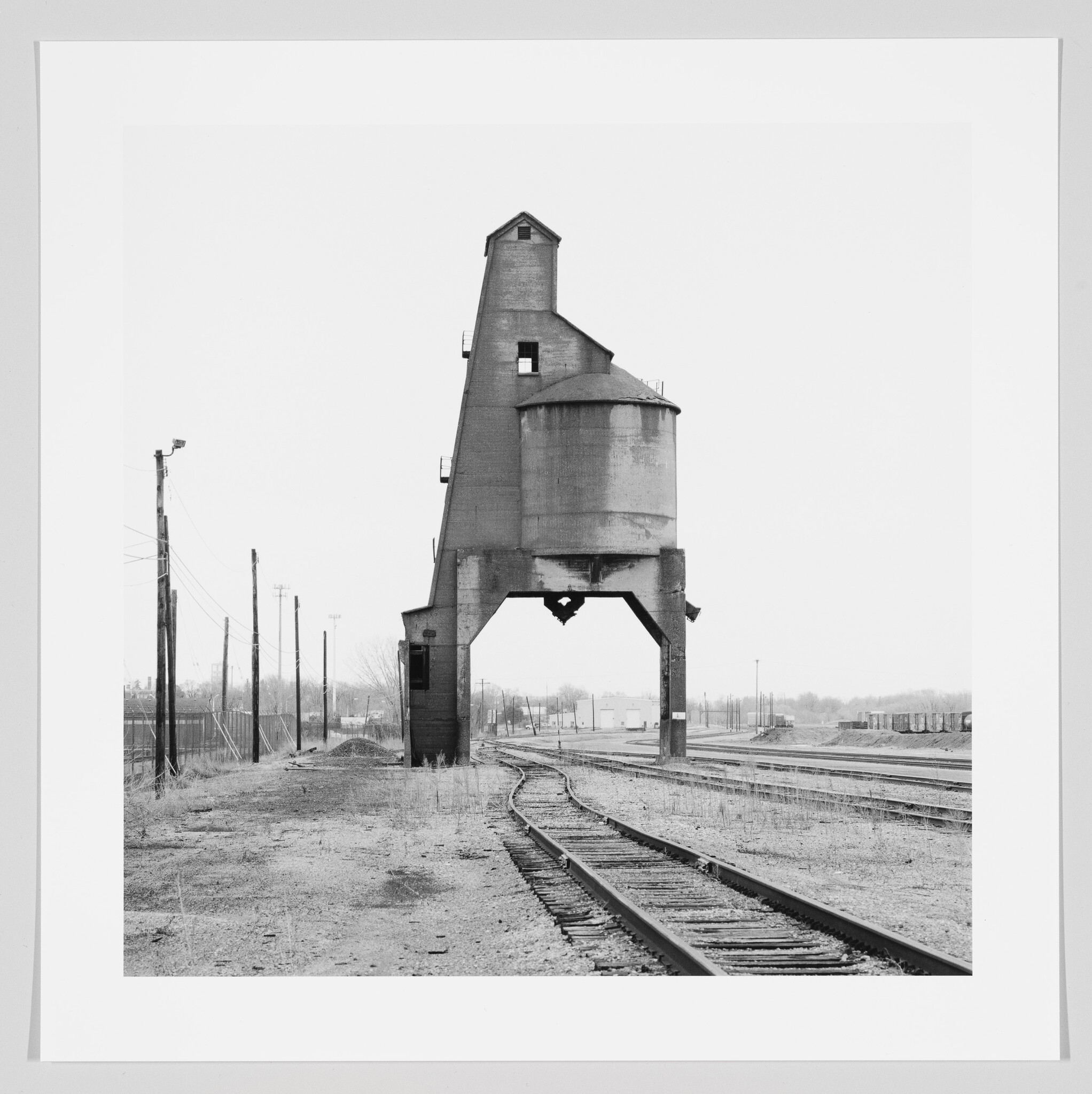 A black and white photograph of an old railroad coaling tower standing alongside multiple railway tracks, with a barren landscape and overcast sky in the background.