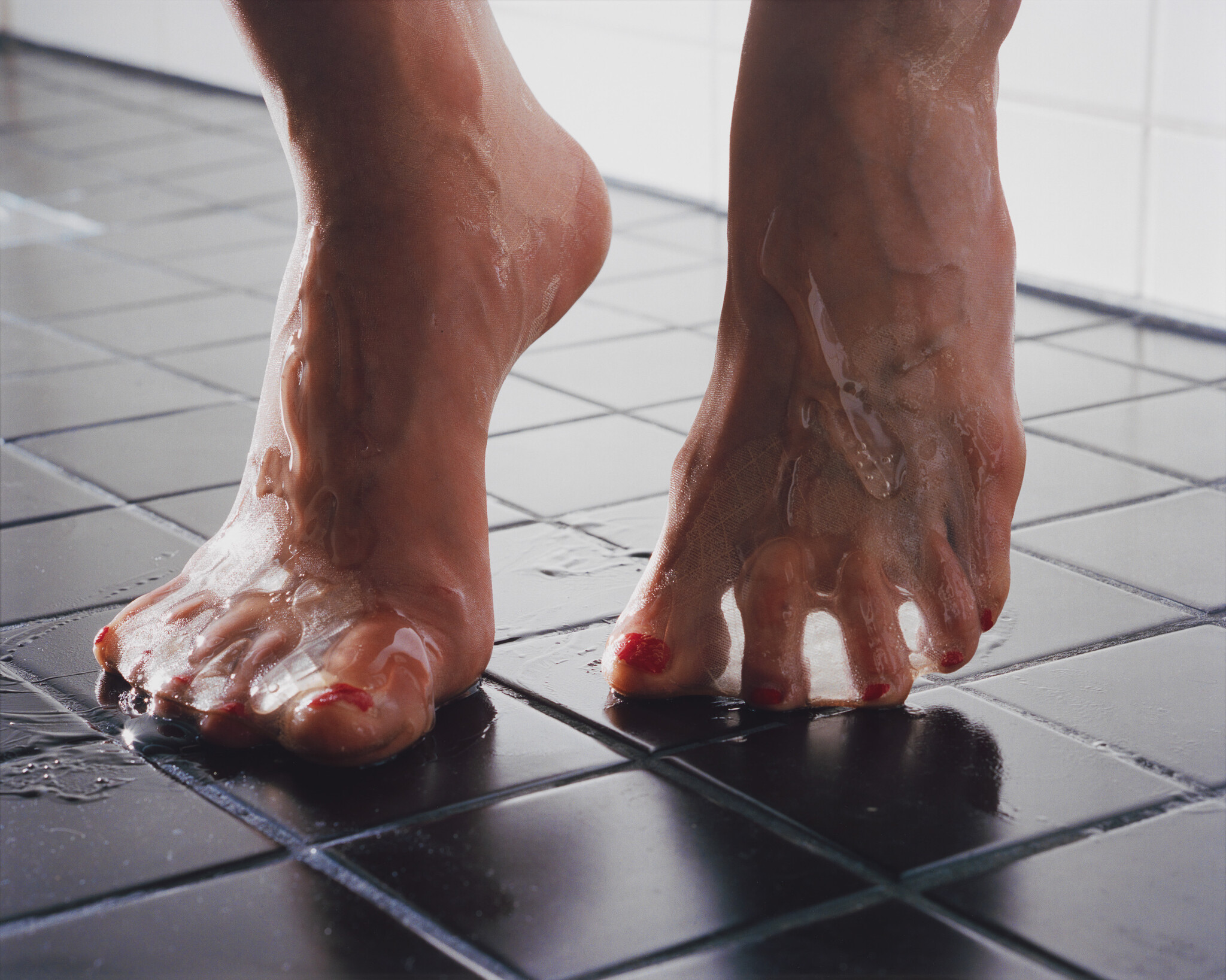 Bare feet with red toenail polish standing on wet black bathroom tiles with water running.
