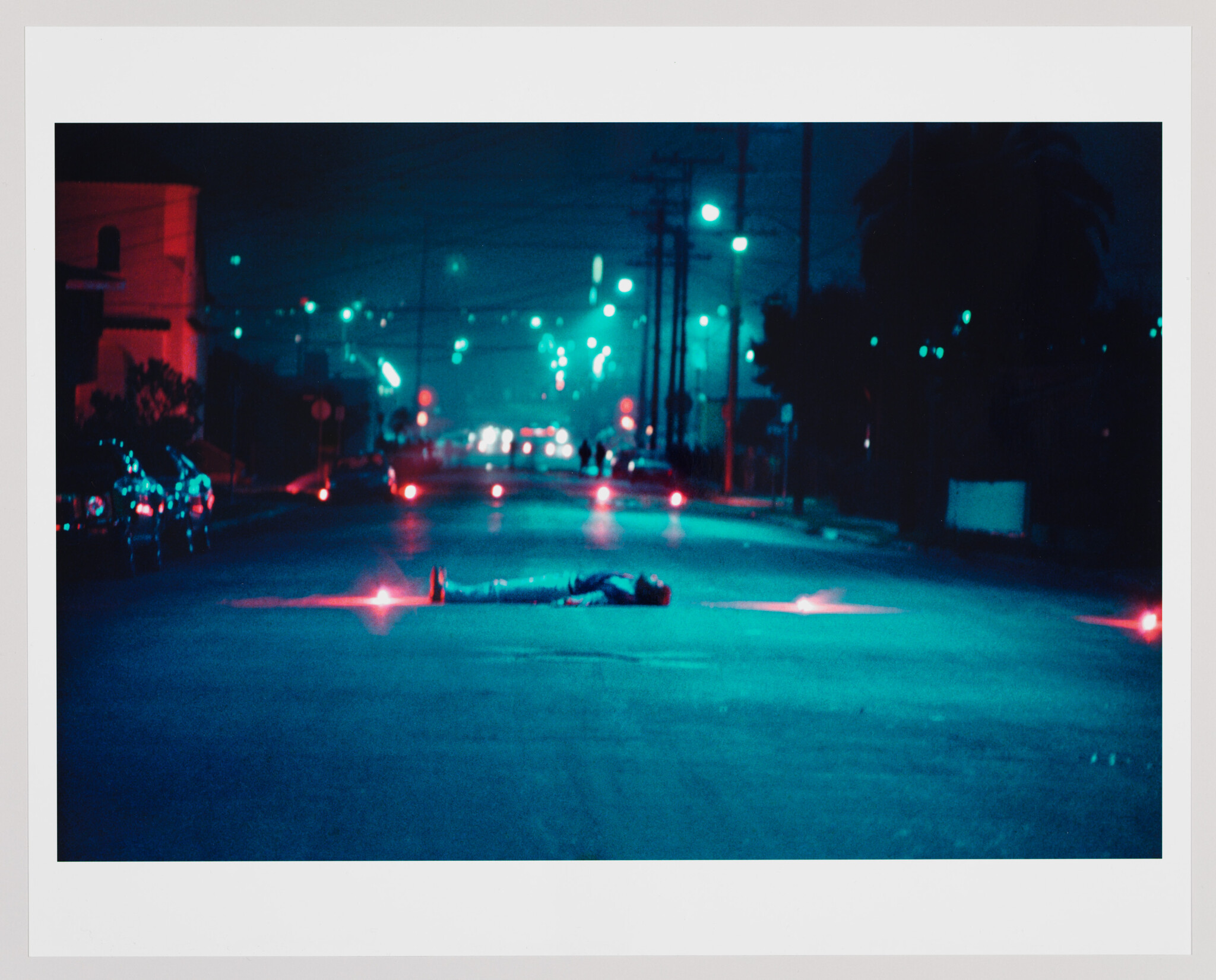 A person lies motionless in the middle of a dim, blue-lit street surrounded by red flares.