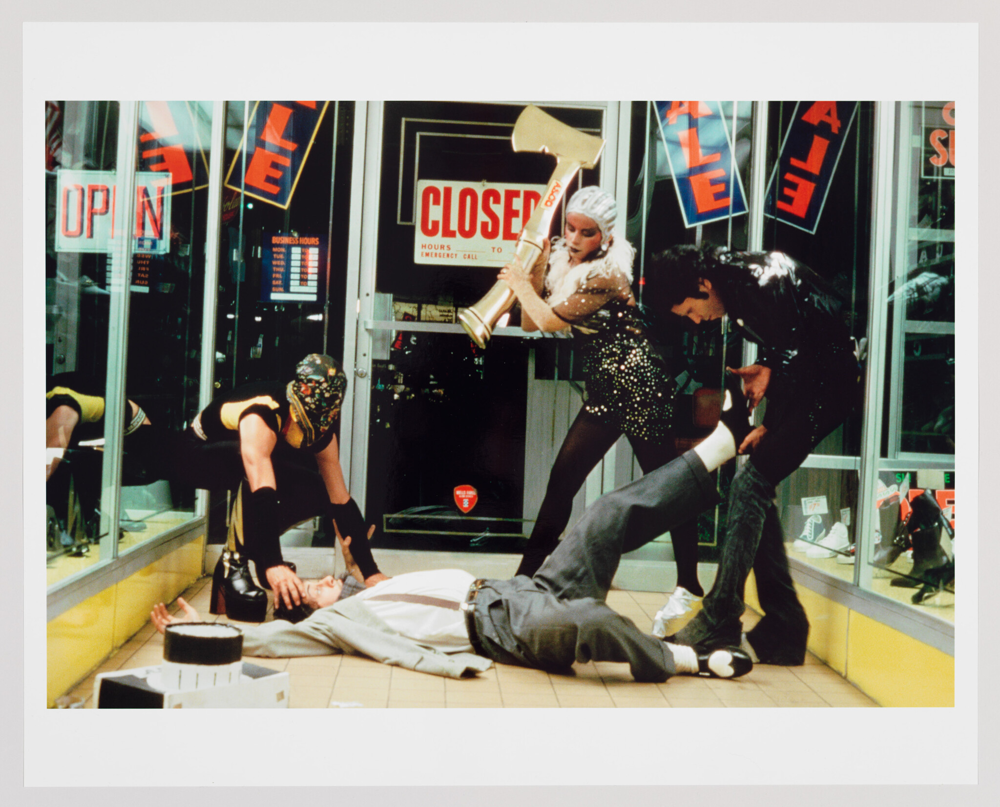A chaotic scene inside a store with "SALE" and "CLOSED" signs on the windows. Three individuals in eccentric costumes and masks appear to be robbing the place, with one person lying on the floor, another holding a saxophone, and the third grabbing a man by the leg. The scene looks like a still from a film or a staged photograph.