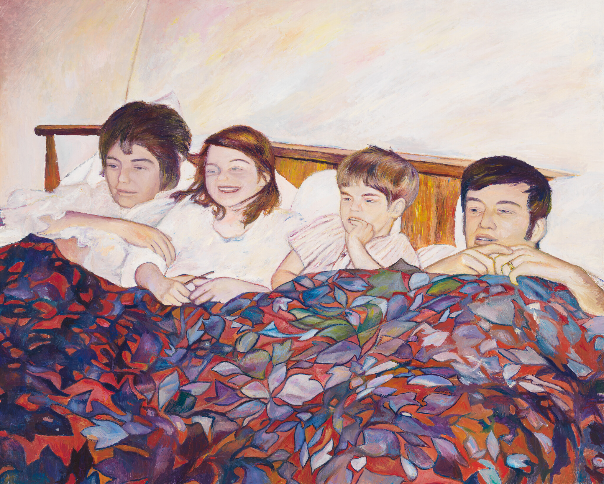 Four family members lying together in bed under a colorful patterned blanket, smiling and relaxed.
