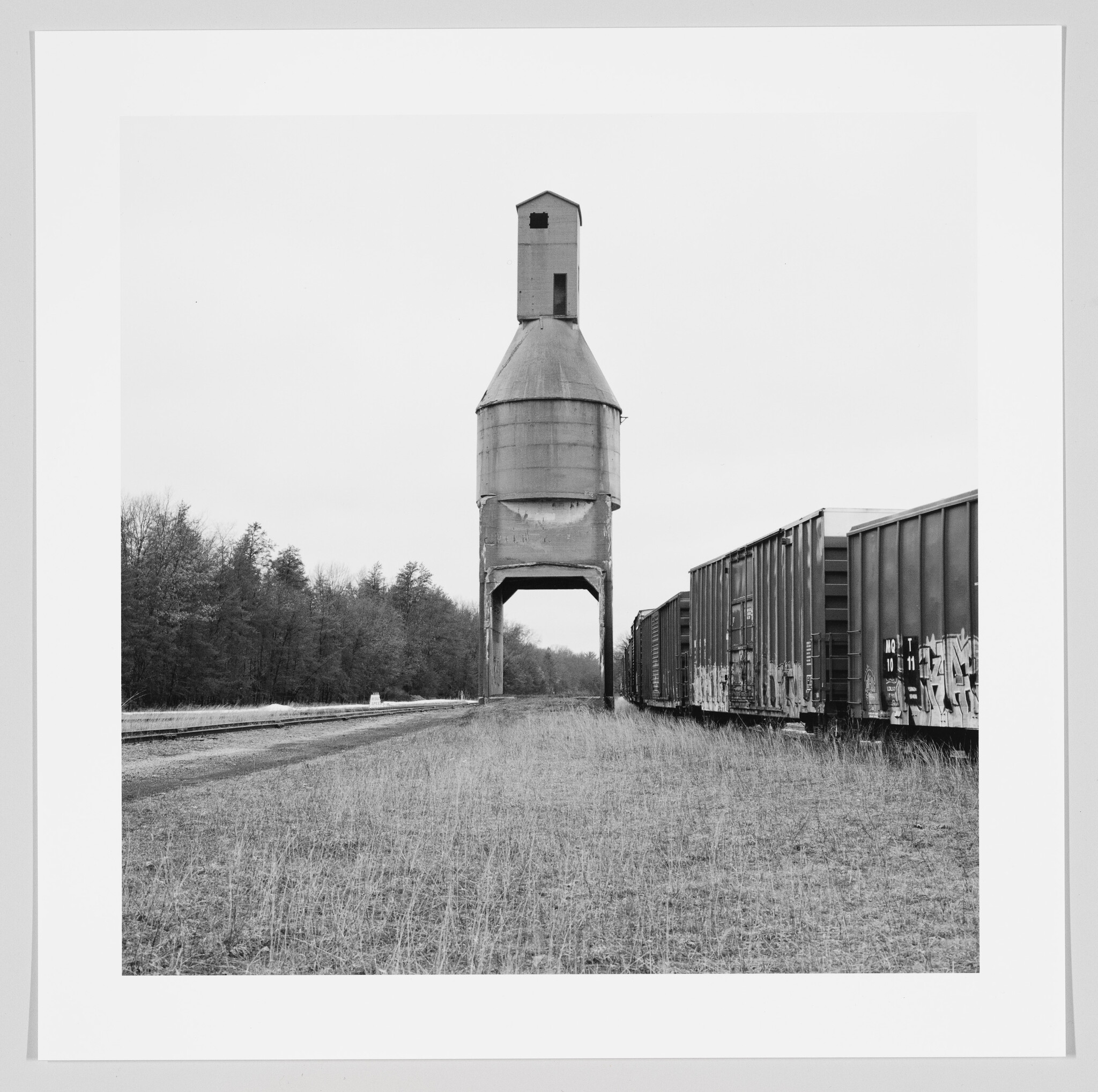 A black and white photograph depicting a vintage railroad water tower standing between two sets of railway tracks, with a freight train parked on the track to the right. The landscape is flat with a sparse tree line in the background, and the overcast sky suggests a gloomy or cold day.