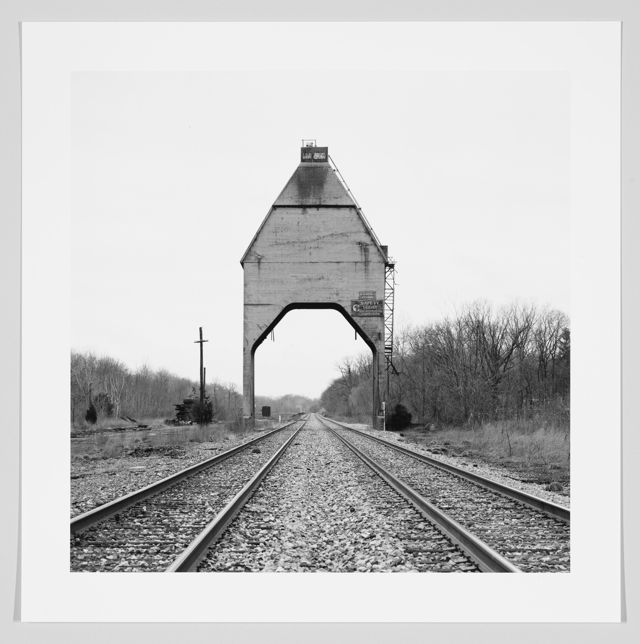 Black and white photograph of a large, historic concrete coaling tower straddling railroad tracks, with barren trees on either side under an overcast sky.