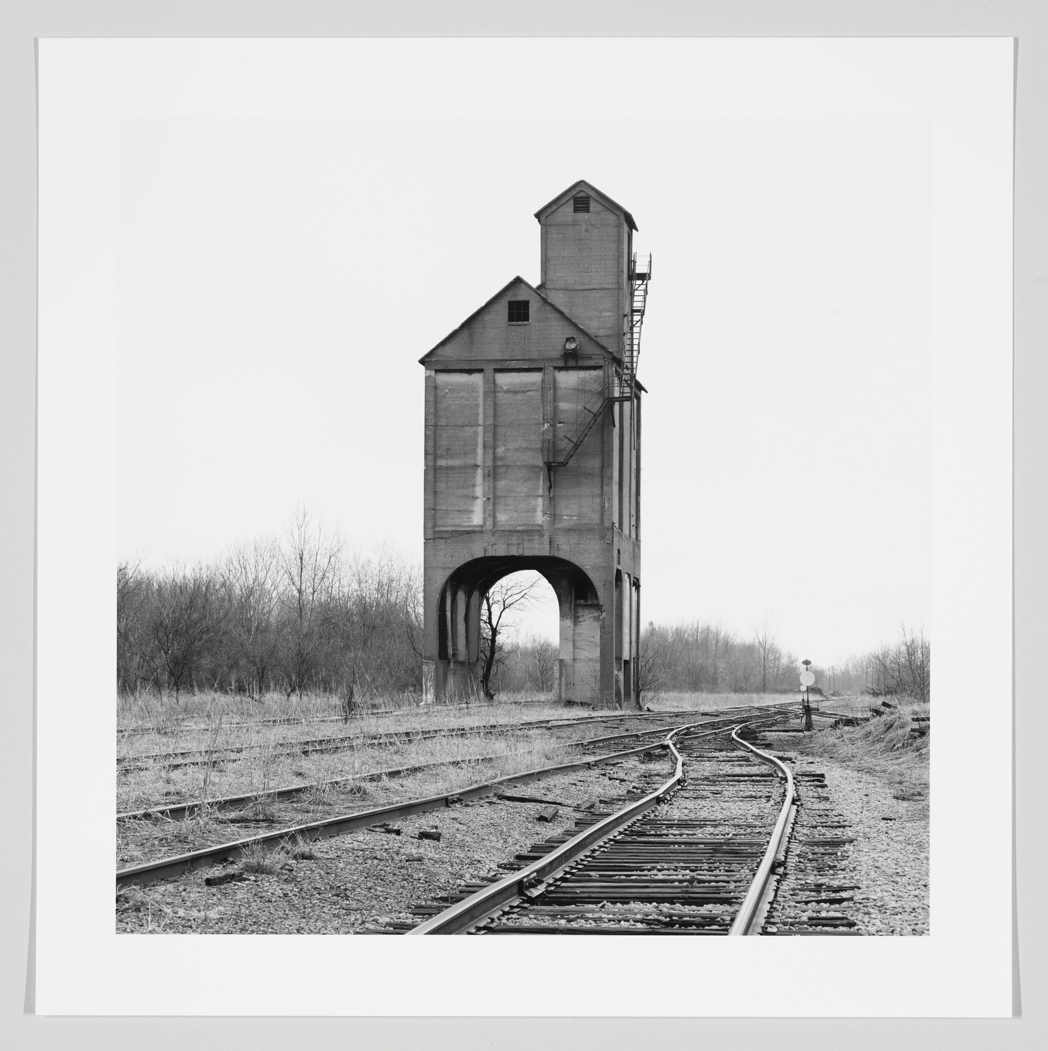 A black and white photograph of an old, tall railroad coaling tower standing over multiple intersecting train tracks, with barren trees in the background and an overcast sky.