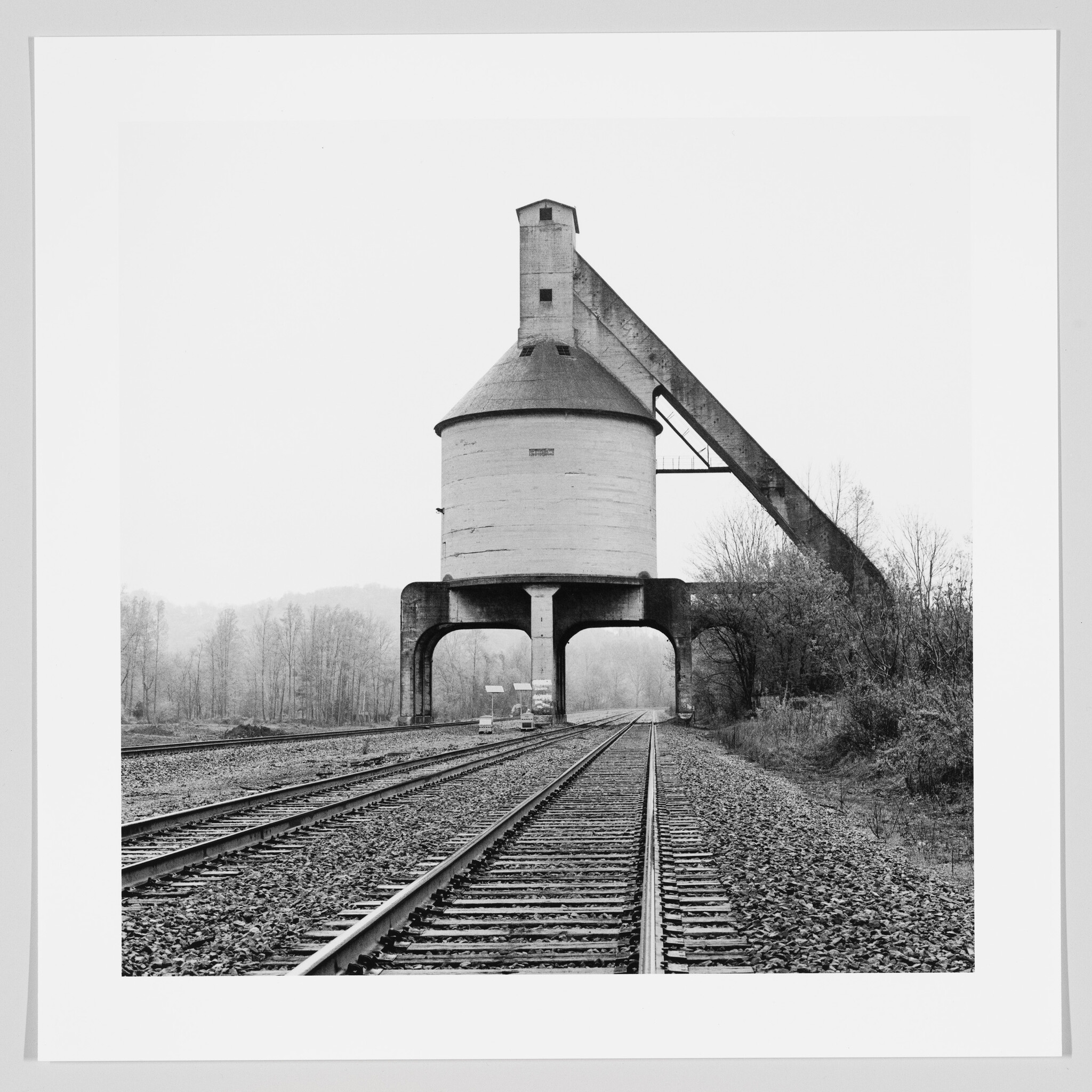 Black and white photograph of a large, old-fashioned concrete coaling tower straddling multiple railroad tracks, with a forested area in the background on an overcast day.
