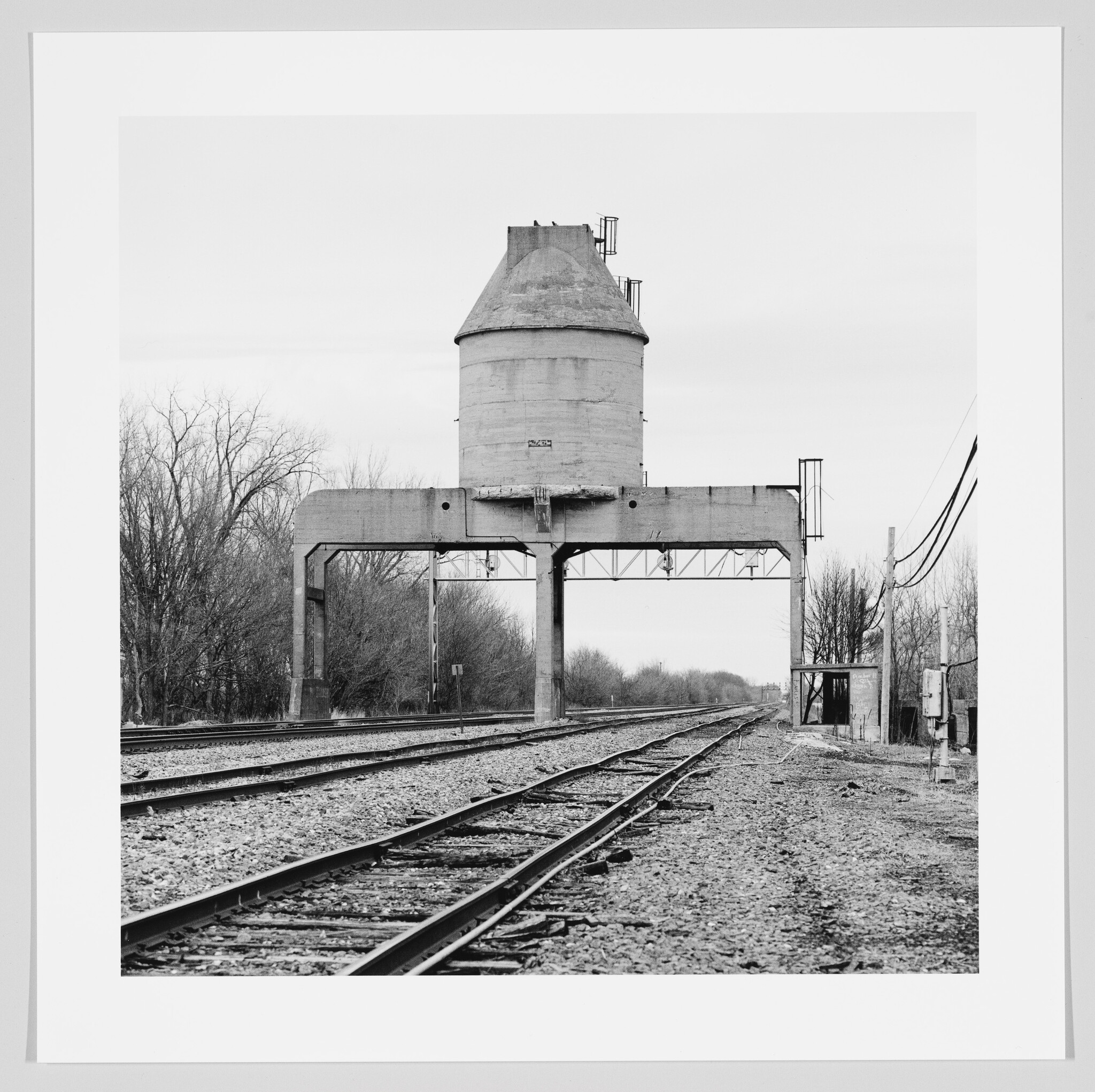 A black and white photograph depicting a vintage concrete coaling tower straddling multiple railroad tracks, with barren trees in the background and a cloudy sky above.