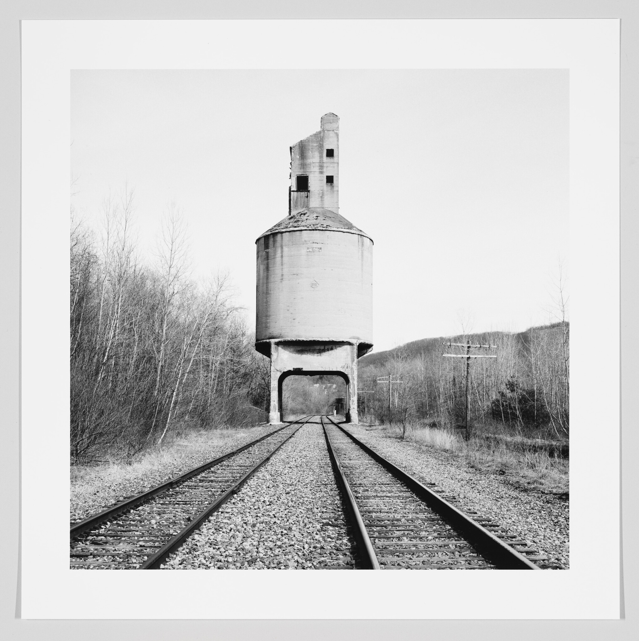 Black and white photograph of a concrete grain elevator towering over railroad tracks, with leafless trees and a hill in the background.