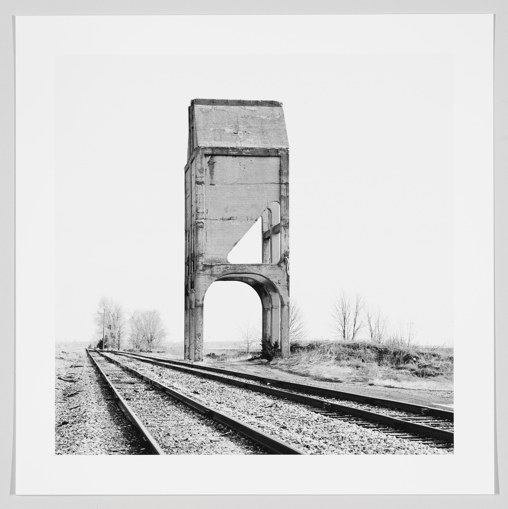 A tall, weathered concrete grain elevator remnant arches over railroad tracks in a flat landscape.