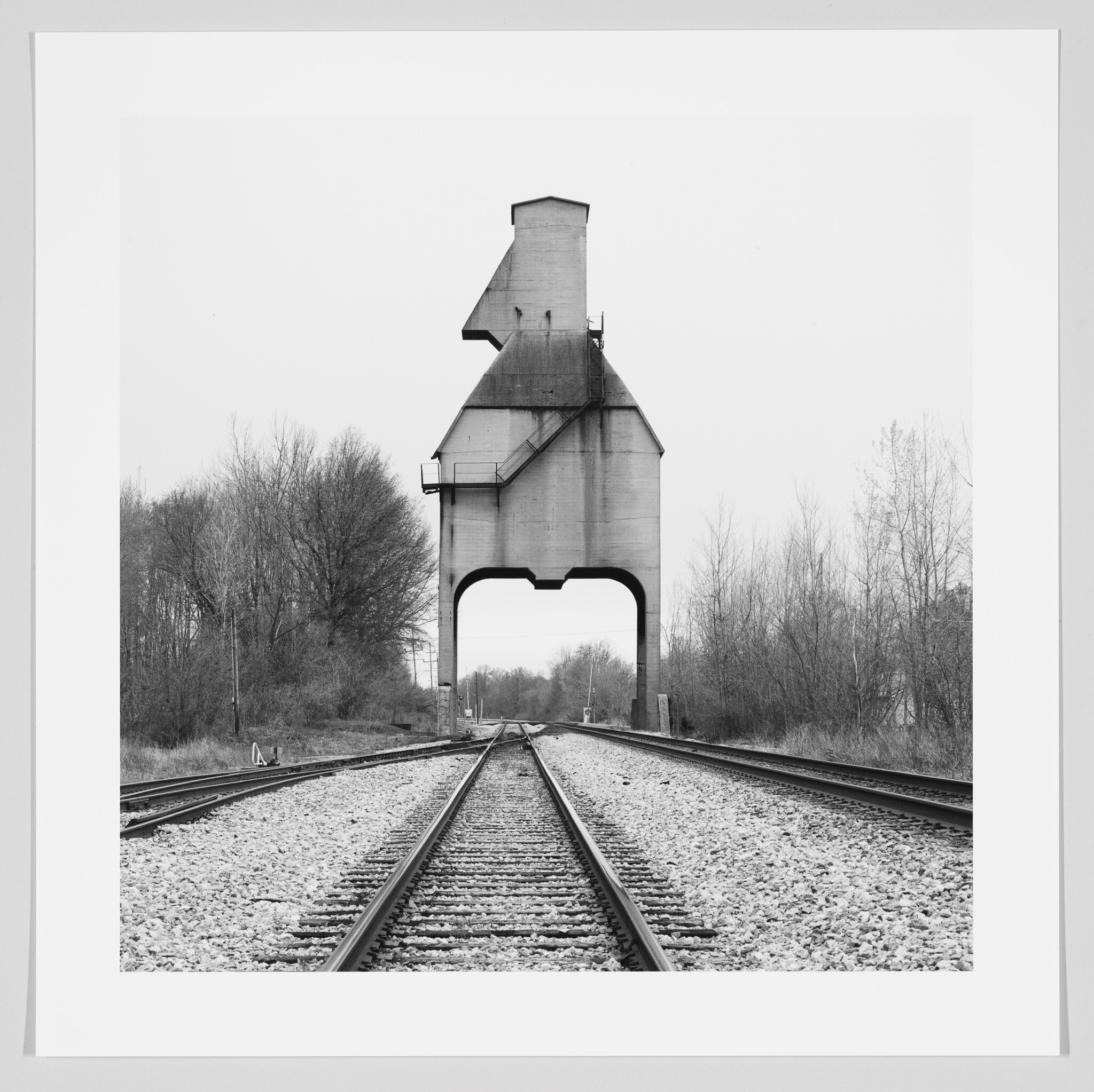 Train tracks lead toward a tall concrete grain elevator spanning the railway in a rural area.