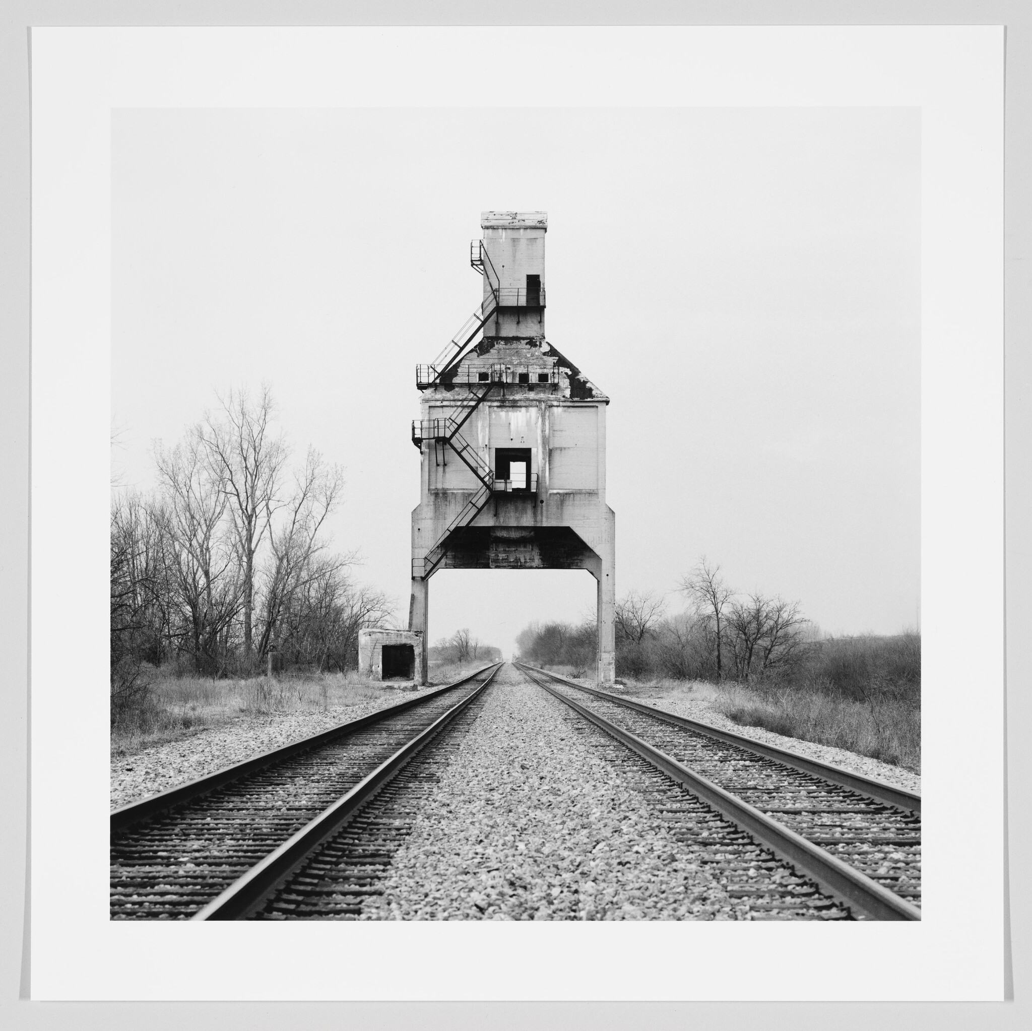 Black and white photograph of a large, old industrial structure straddling railroad tracks, with barren trees in the background under an overcast sky.