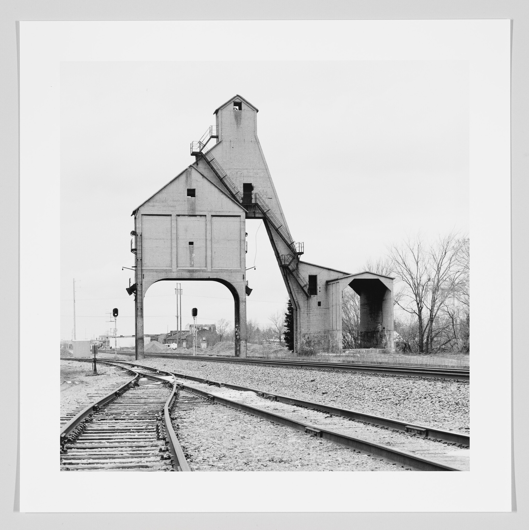 A black and white photograph of a vintage coal chute structure towering over railroad tracks, with multiple lines converging in the foreground and bare trees in the background under an overcast sky.
