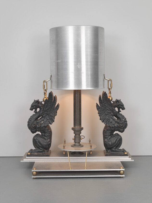 A decorative table lamp with two griffin sculptures supporting a central metal shade and lit bulb.
