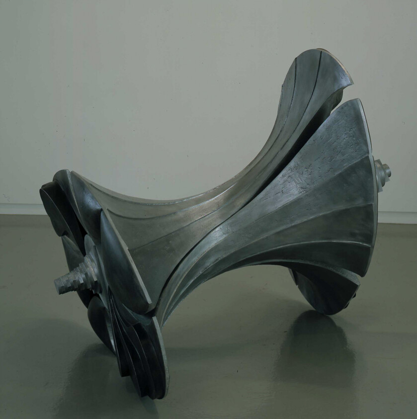A large twisted metal sculpture resembling a double turbine rotor resting on a gallery floor.