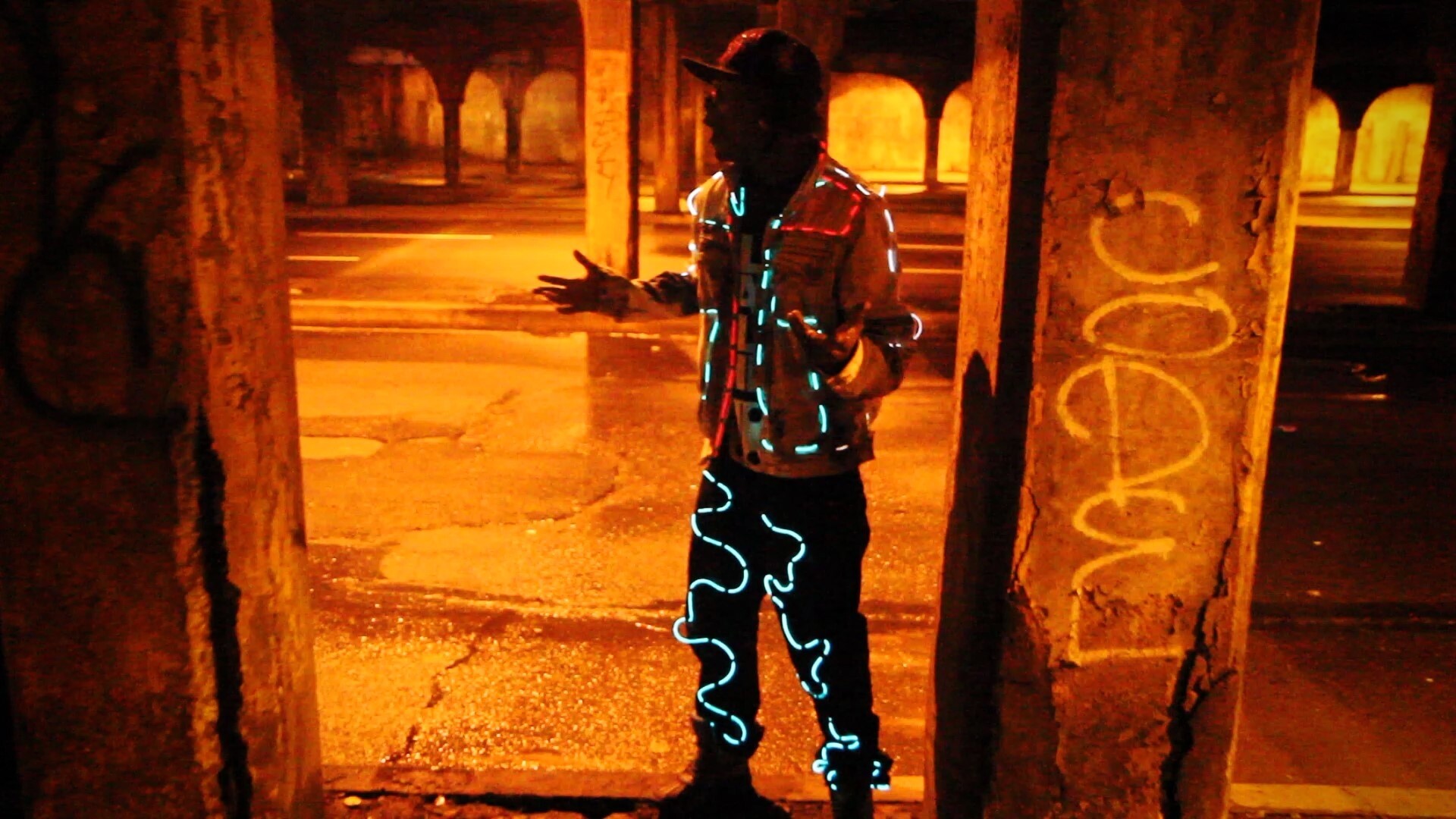 Person wearing glowing, light-up clothing stands under a bridge at night, with graffiti on a nearby pillar.