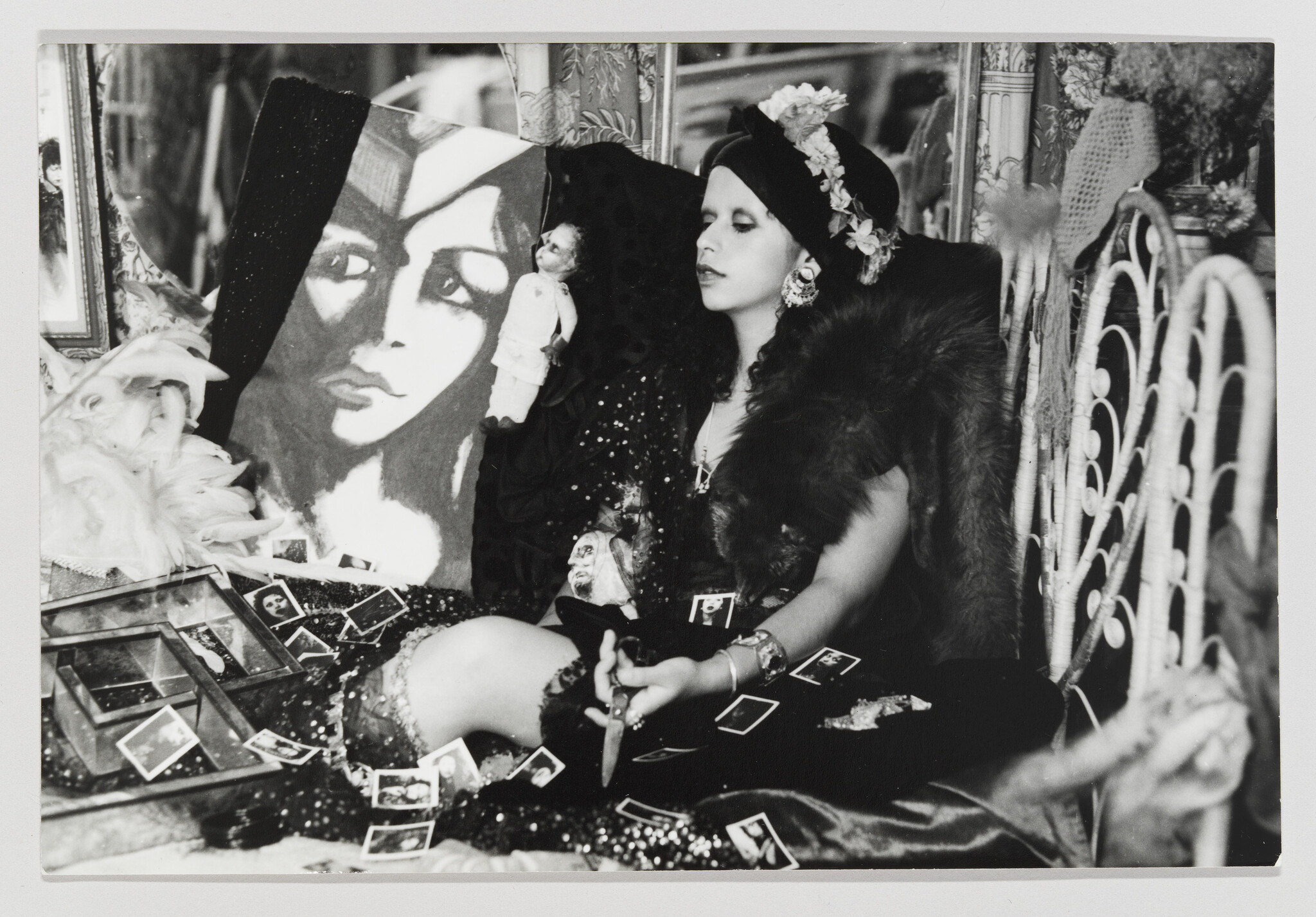 A woman in a turban and fur holds a dagger while surrounded by photos and a painted portrait.