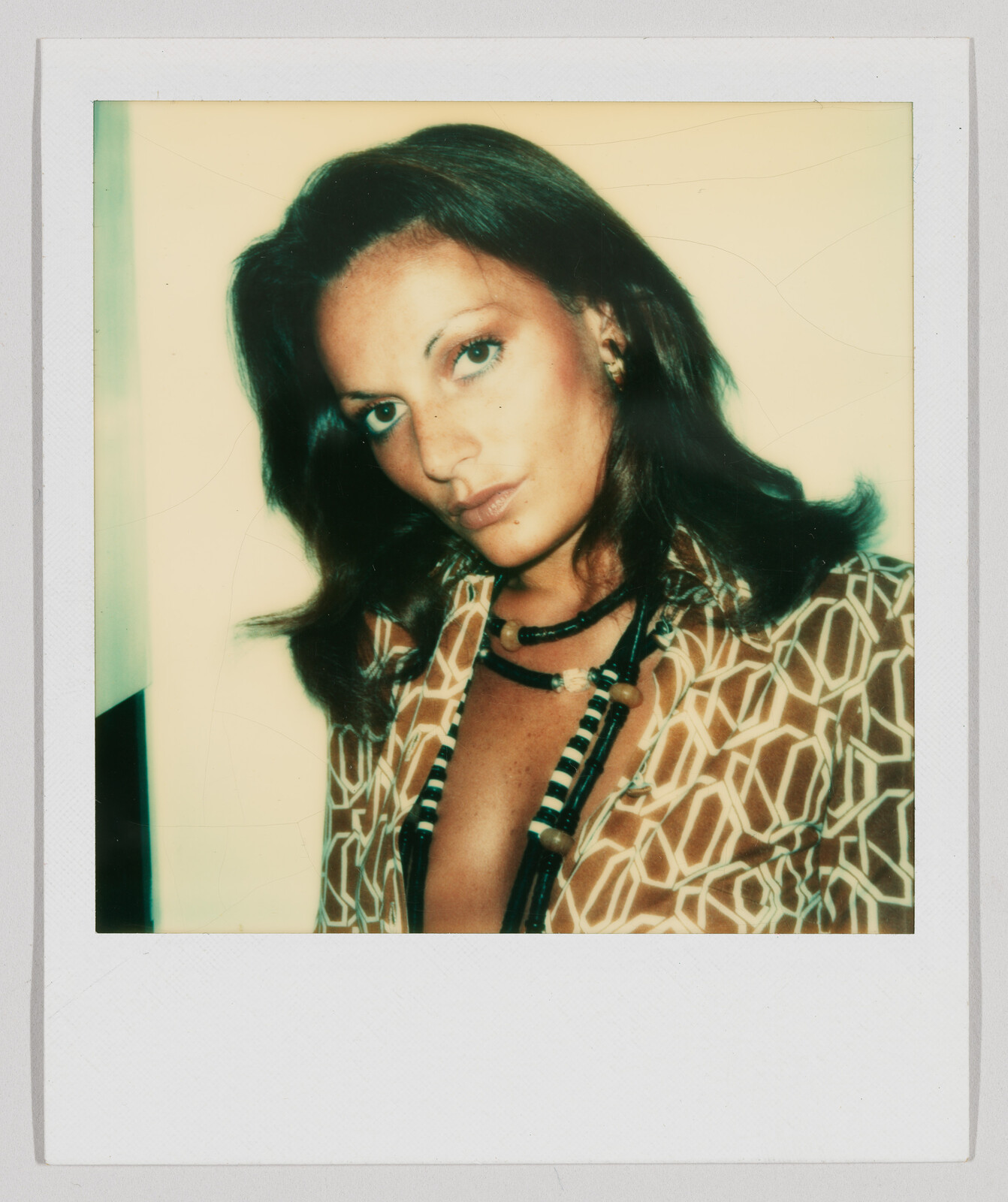 Woman with shoulder-length dark hair wearing a patterned shirt and layered necklaces, looking directly at the camera.