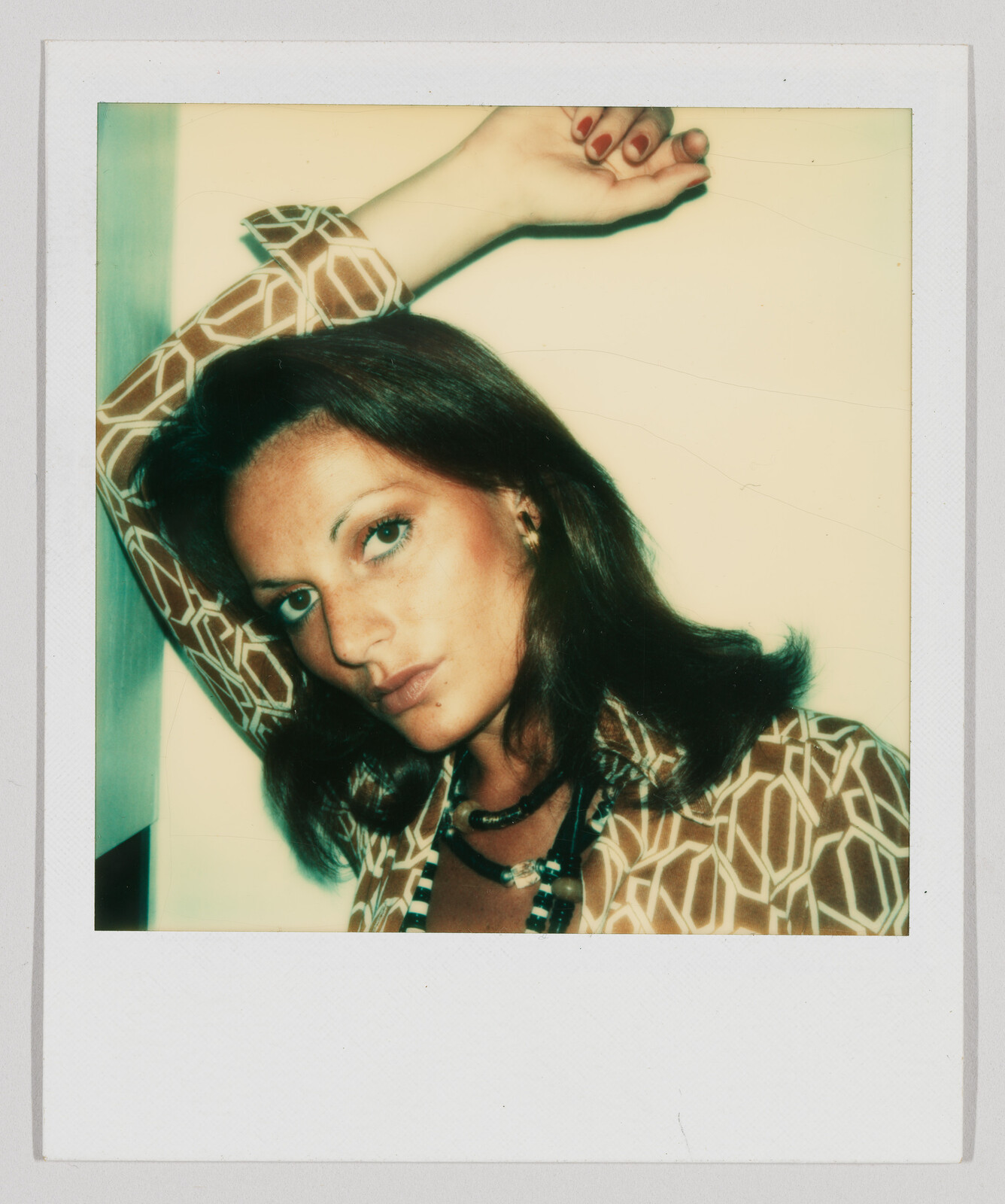 Woman with brown hair leans against a wall, wearing a patterned shirt and necklace, arm raised overhead.