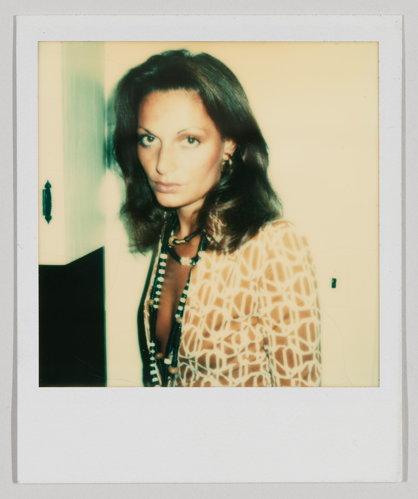 Woman with shoulder-length brown hair wearing a patterned top and long beaded necklace, standing indoors.