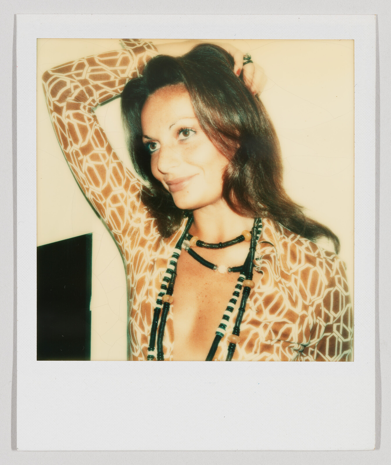 Woman with long brown hair smiles, wearing a patterned shirt and beaded necklace, with one arm raised.