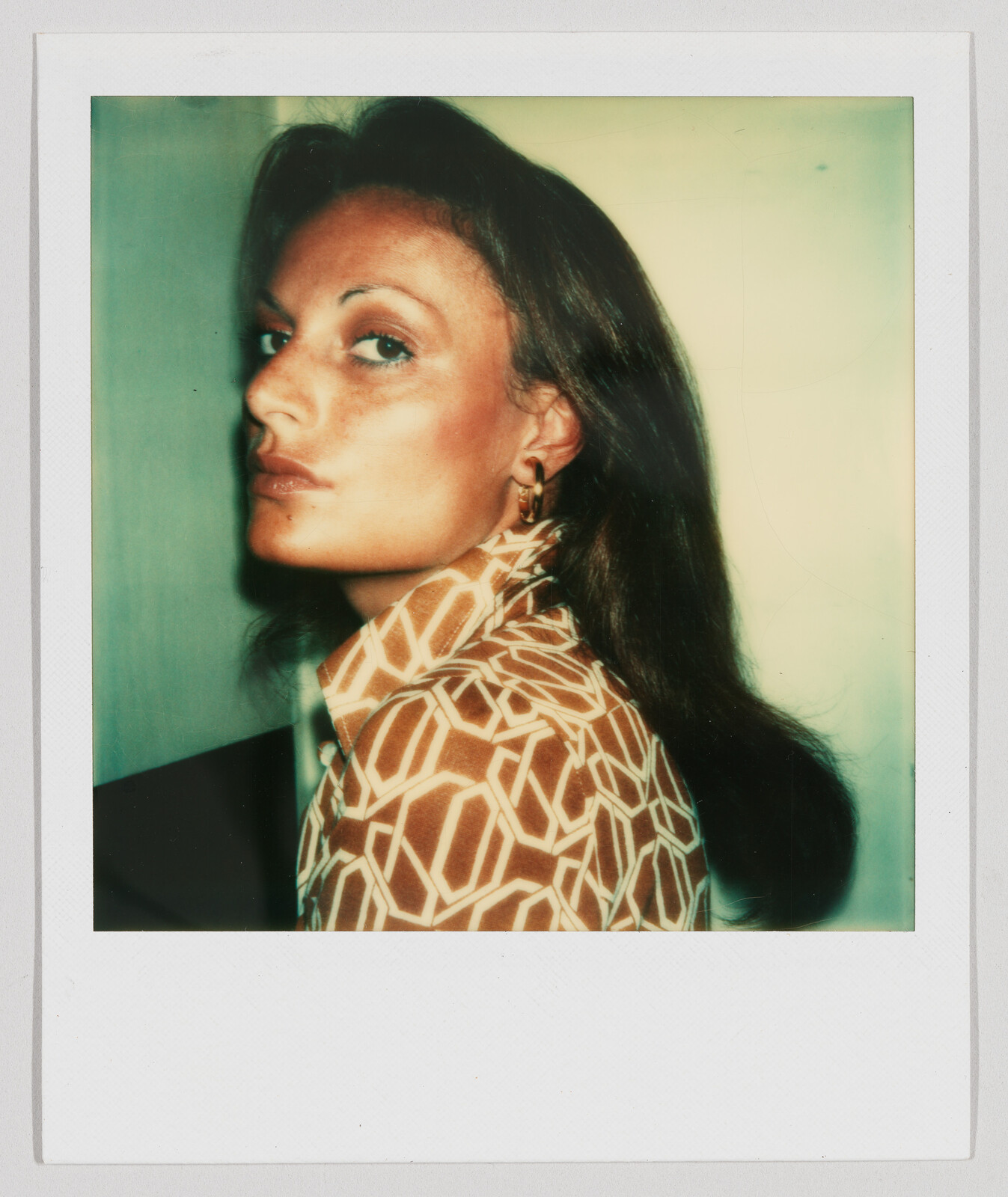 Woman with long brown hair wearing a patterned shirt looks over her shoulder with a neutral expression.