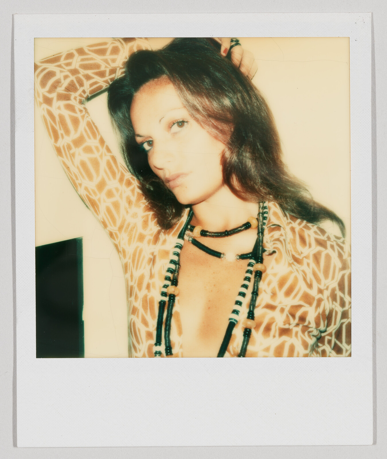 Woman with long brown hair wearing a patterned shirt and beaded necklaces, posing with one arm raised.