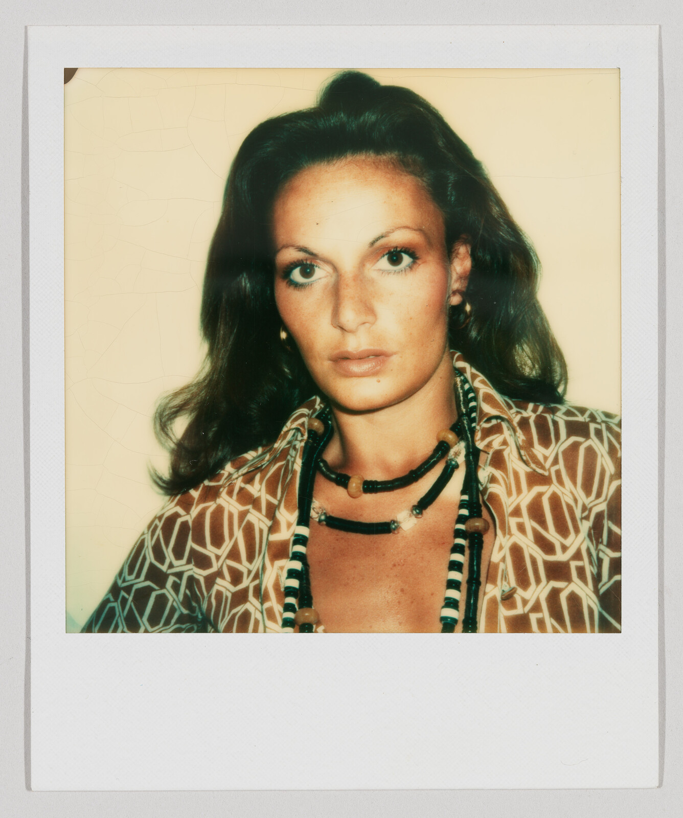 Woman with long brown hair wearing a patterned shirt and layered necklaces, looking directly at the camera.