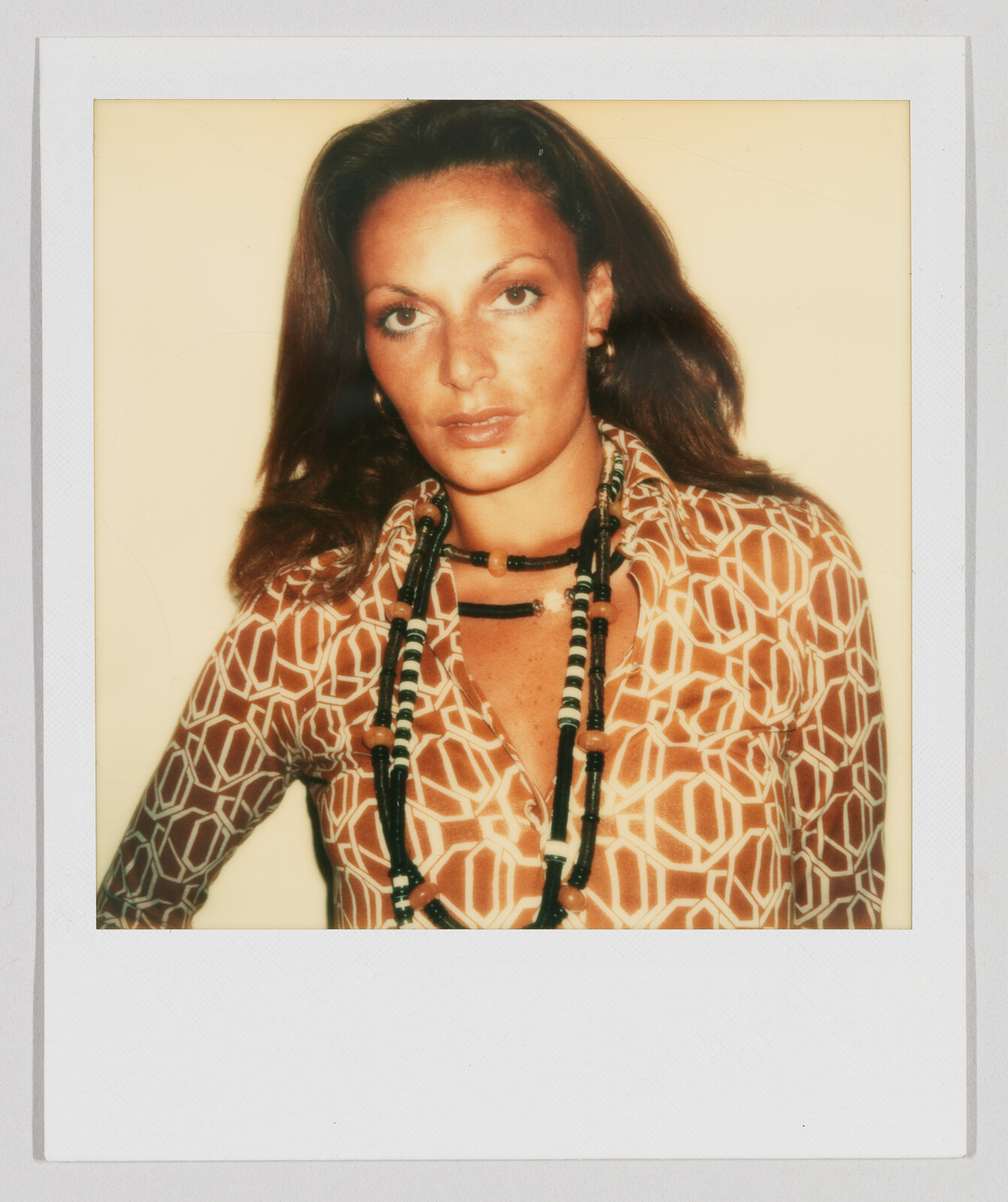 Woman with long brown hair wearing a patterned shirt and long beaded necklaces, looking directly at the camera.