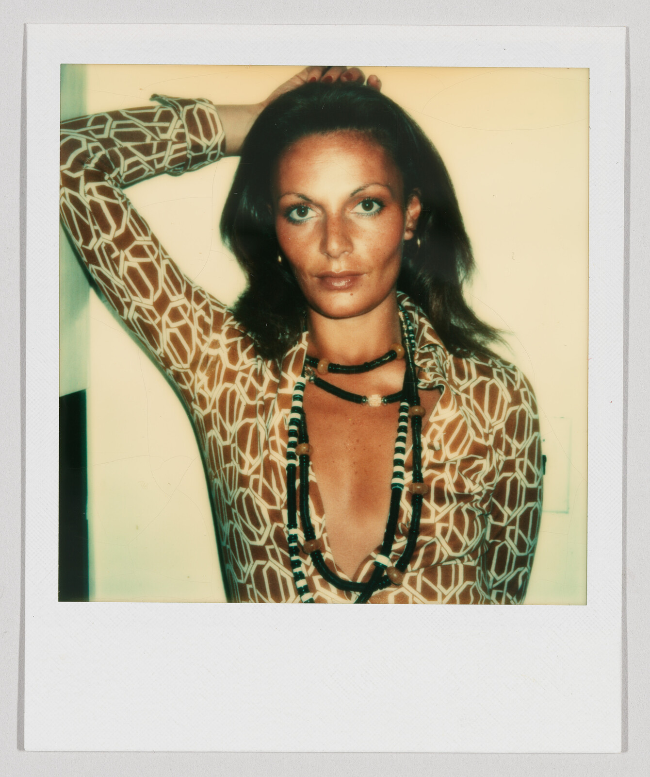 Woman with long dark hair wearing a patterned shirt and layered necklaces, resting one arm on her head.