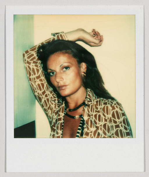 Woman with patterned blouse and necklaces poses with one arm raised behind her head.
