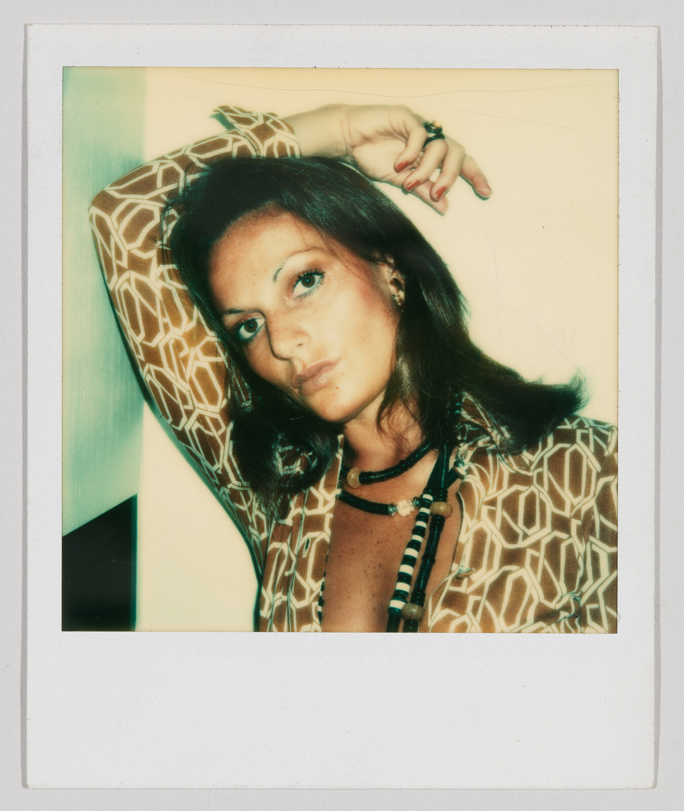 Woman with brown hair poses with one arm raised, wearing a patterned shirt and beaded necklaces.