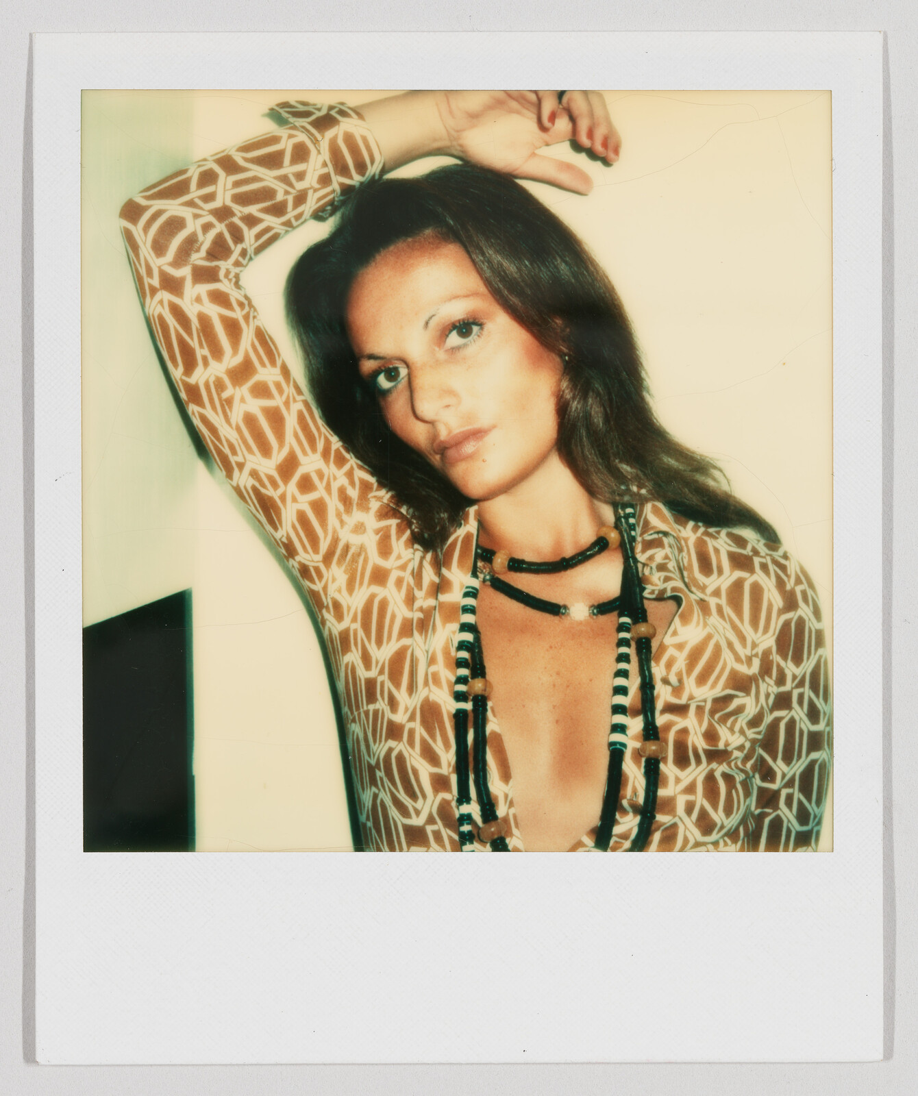 Woman with long brown hair wearing a patterned shirt and beaded necklaces, resting one arm on her head.