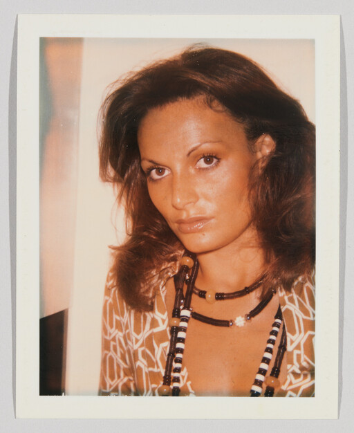 Woman with shoulder-length brown hair wearing patterned blouse and layered beaded necklaces, looking at camera.
