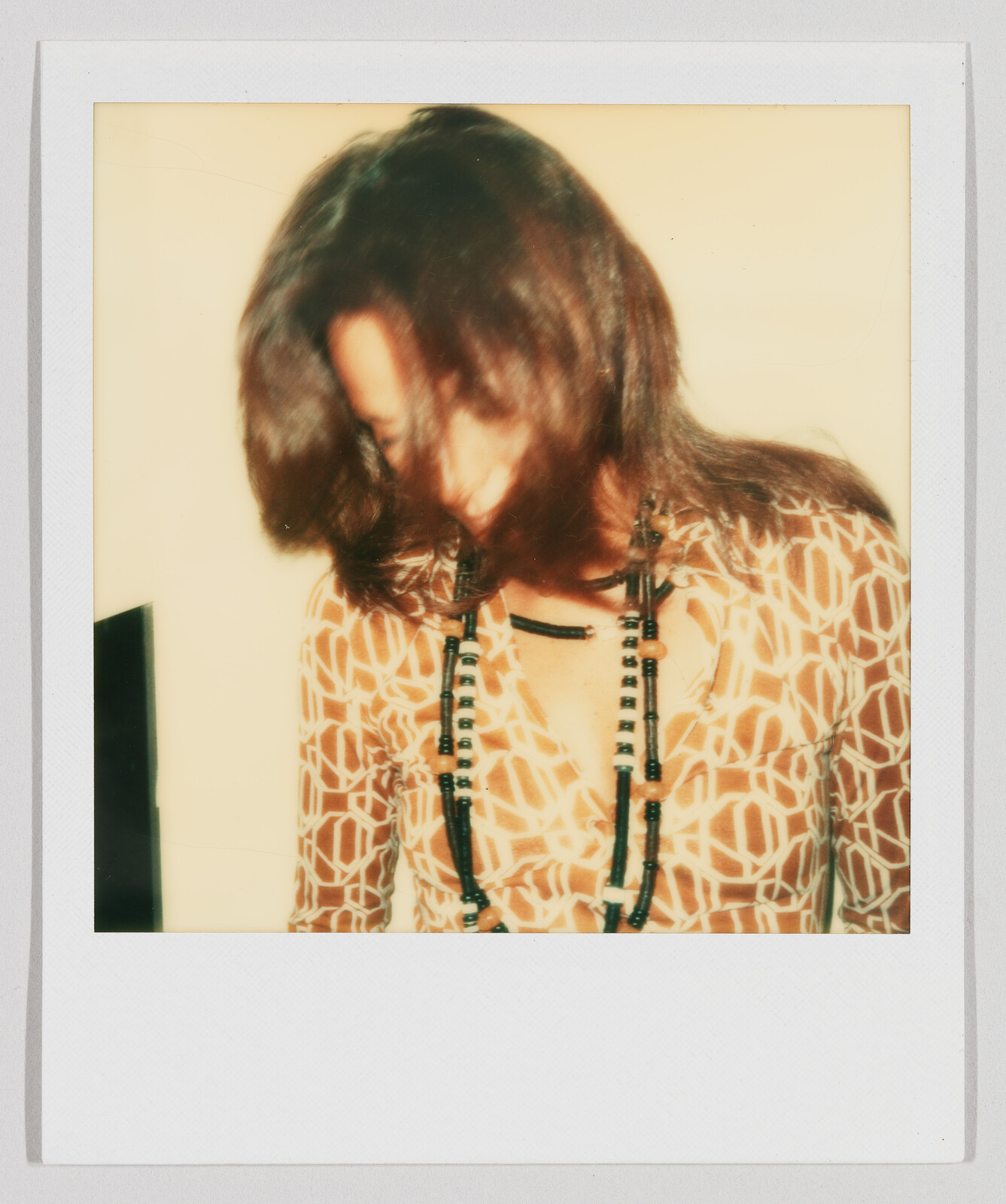 Person with long brown hair looks down, wearing a patterned shirt and a long beaded necklace.