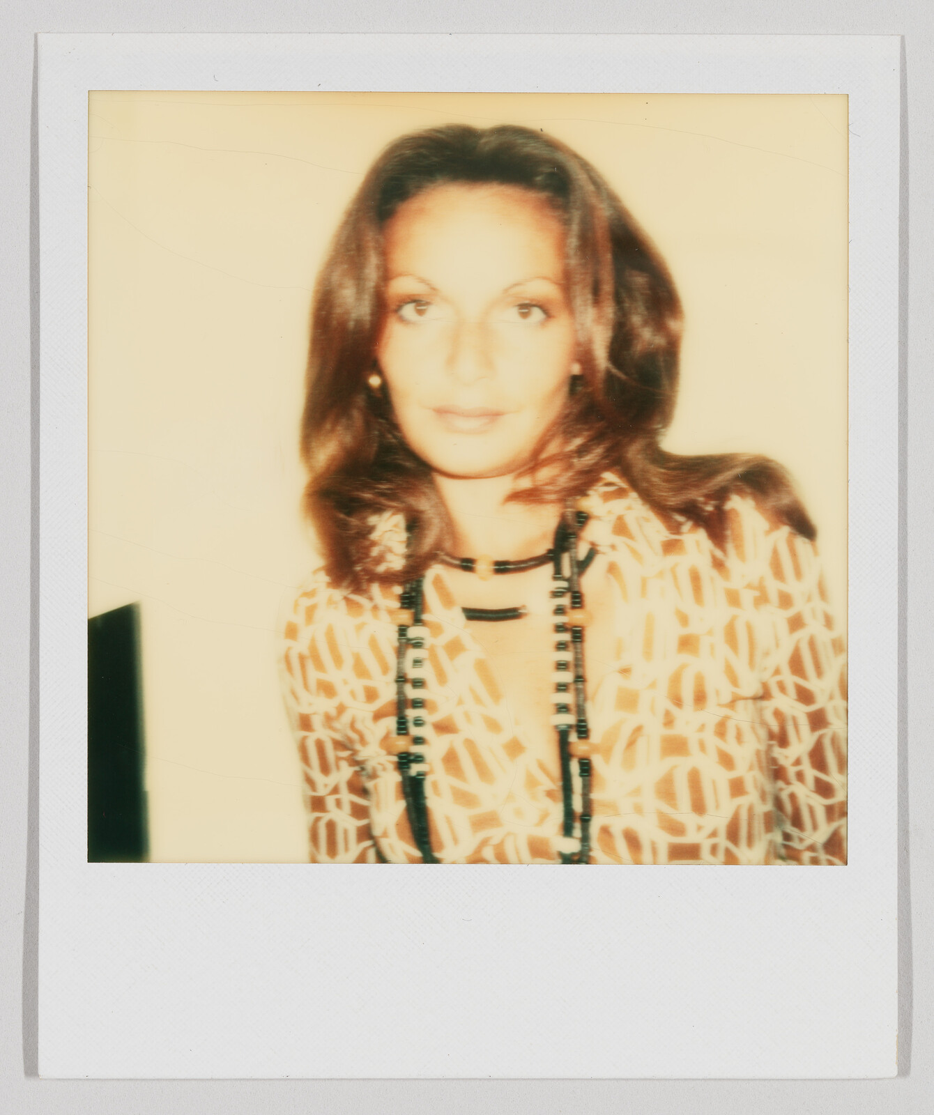 Woman with long brown hair wearing a patterned shirt and a long beaded necklace, looking at the camera.