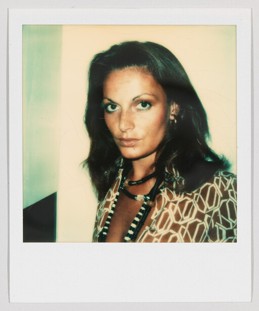A woman with dark hair wearing a geometric patterned blouse and layered beaded necklaces looks toward the camera.