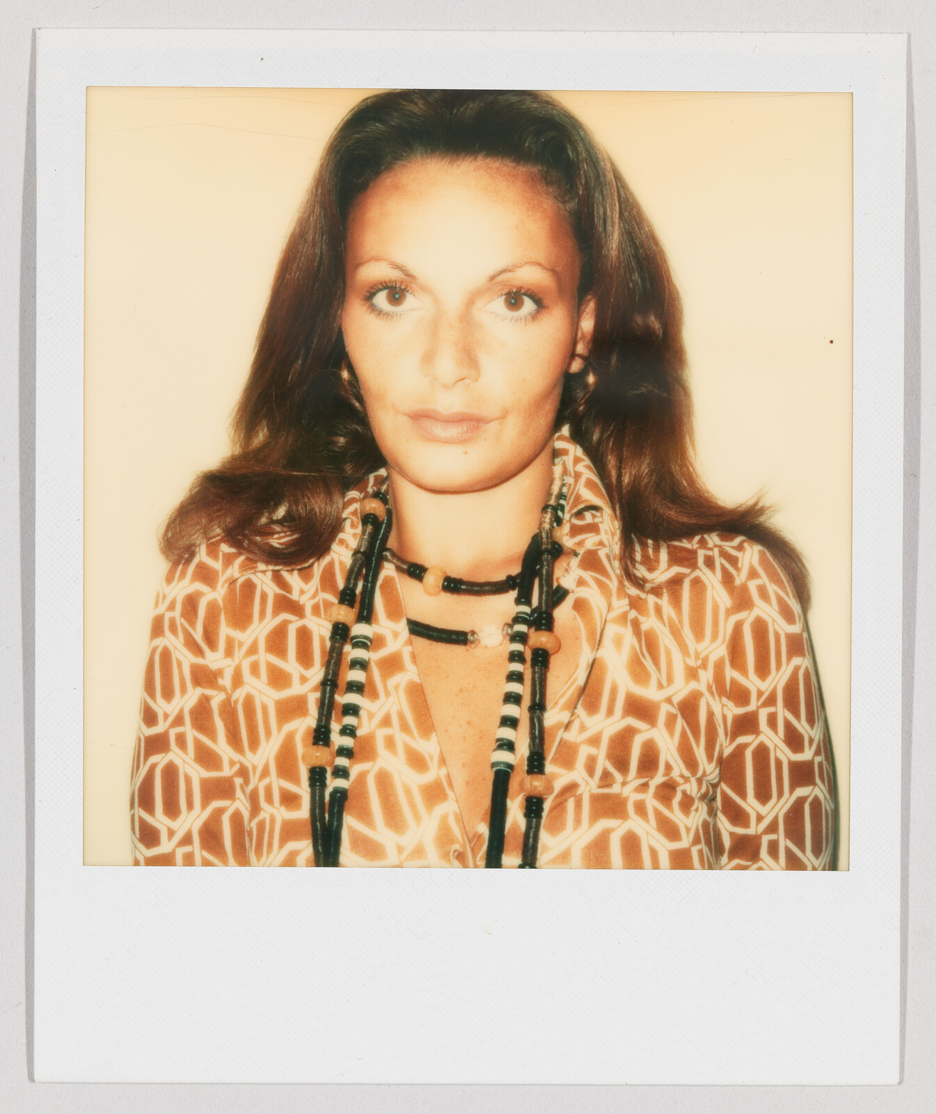 Woman with long brown hair wearing a patterned shirt and layered beaded necklaces, looking directly at the camera.