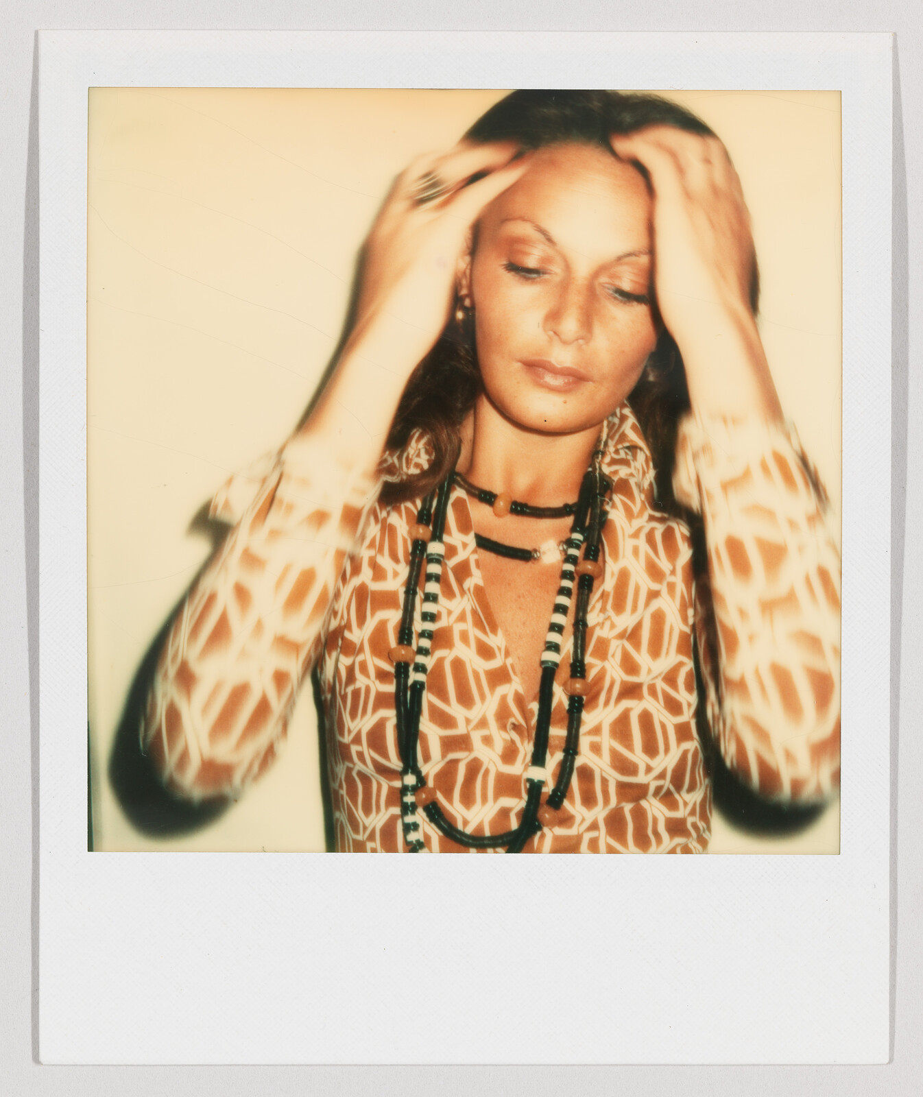 Woman with long hair and patterned shirt touches her head, wearing layered necklaces and looking down.