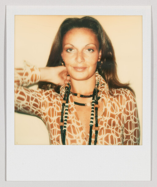 A woman wearing a patterned blouse and layered beaded necklaces looks at the camera with her hand behind her neck.