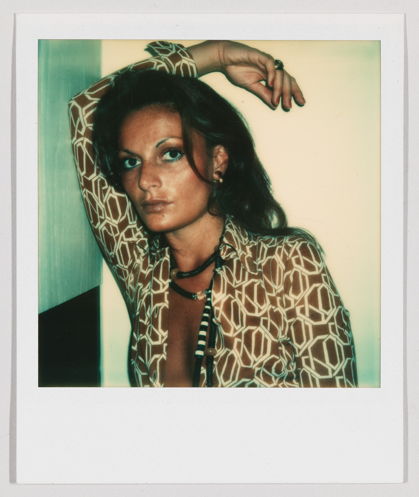 Woman with long brown hair wearing a patterned shirt, leaning against a wall with one arm raised.