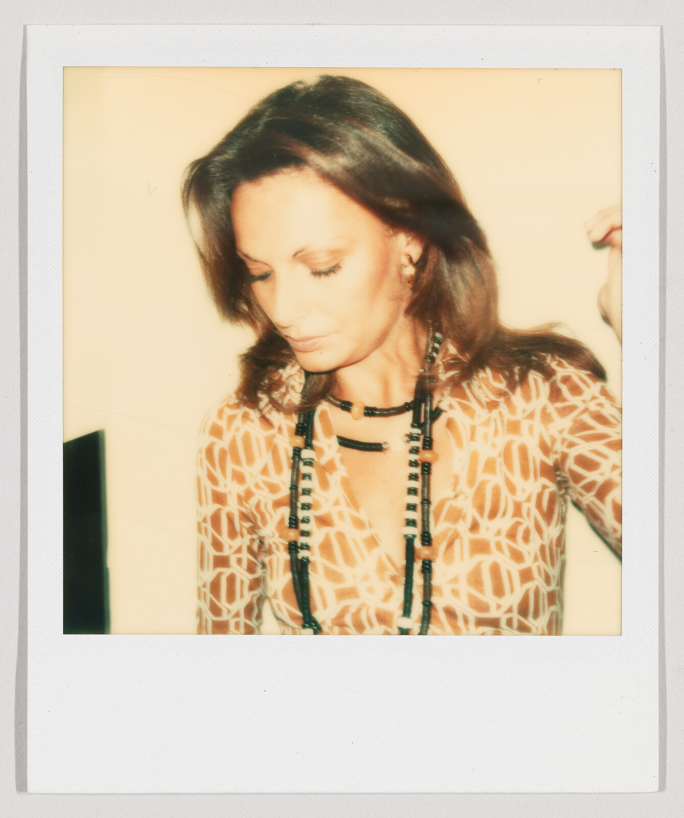 Woman with shoulder-length brown hair looks down, wearing a patterned shirt and long black beaded necklace.