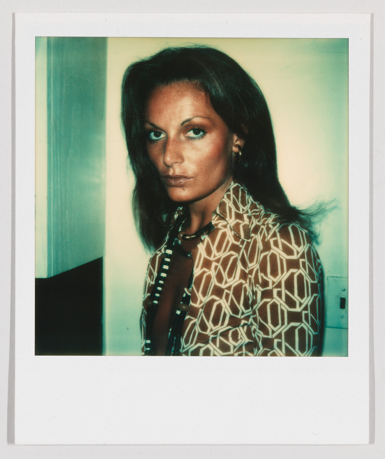 Woman with long brown hair wearing a patterned shirt looks toward the camera with a neutral expression.