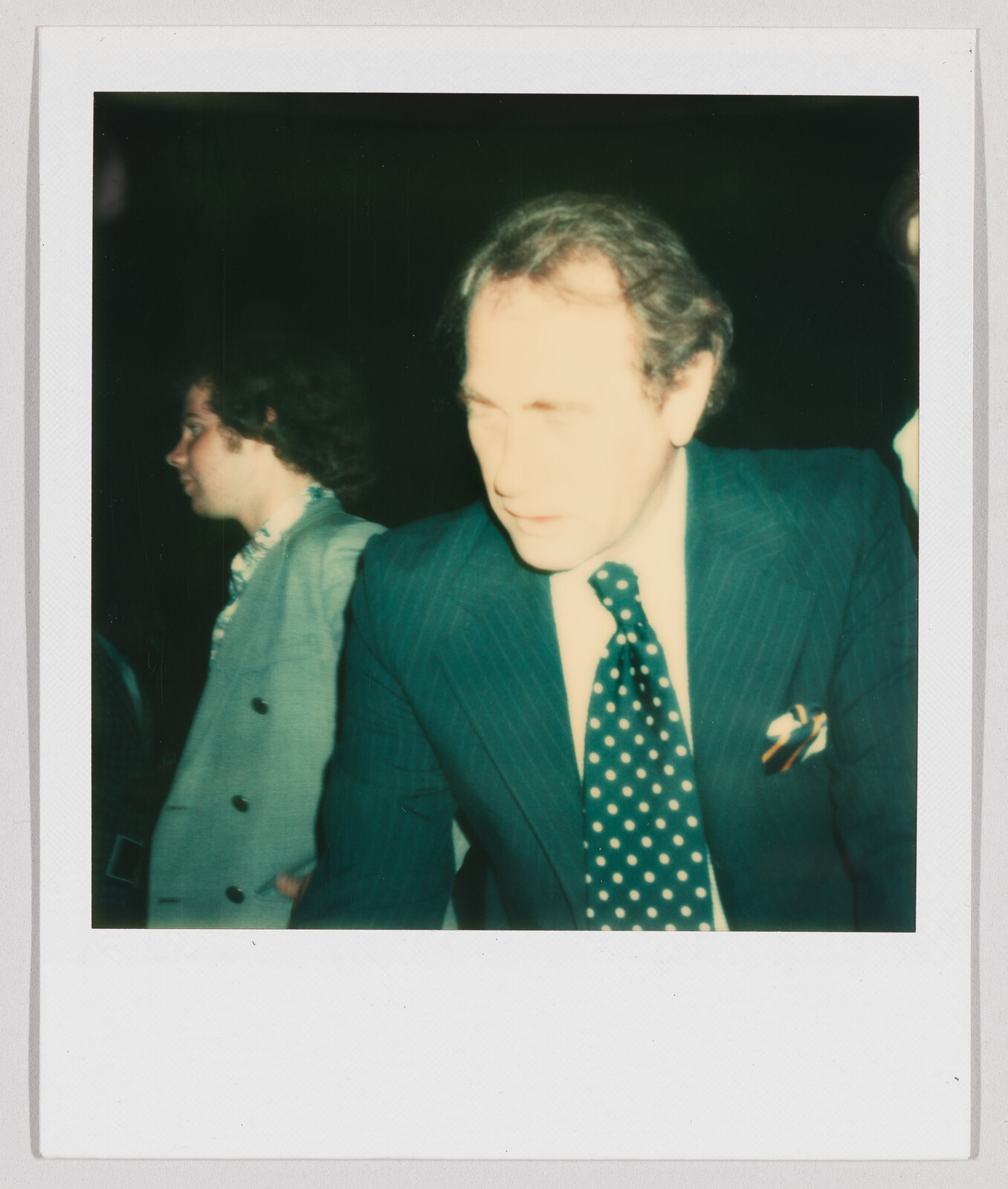 Two men in suits stand indoors, one in front with a polka dot tie, both looking away from the camera.