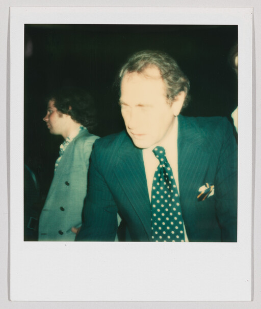 A middle-aged man in a striped suit and polka-dot tie leans forward at a dim event.