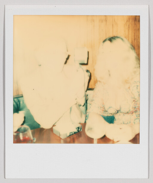 Two people sit at a table in a bright, overexposed Polaroid holding drinks.