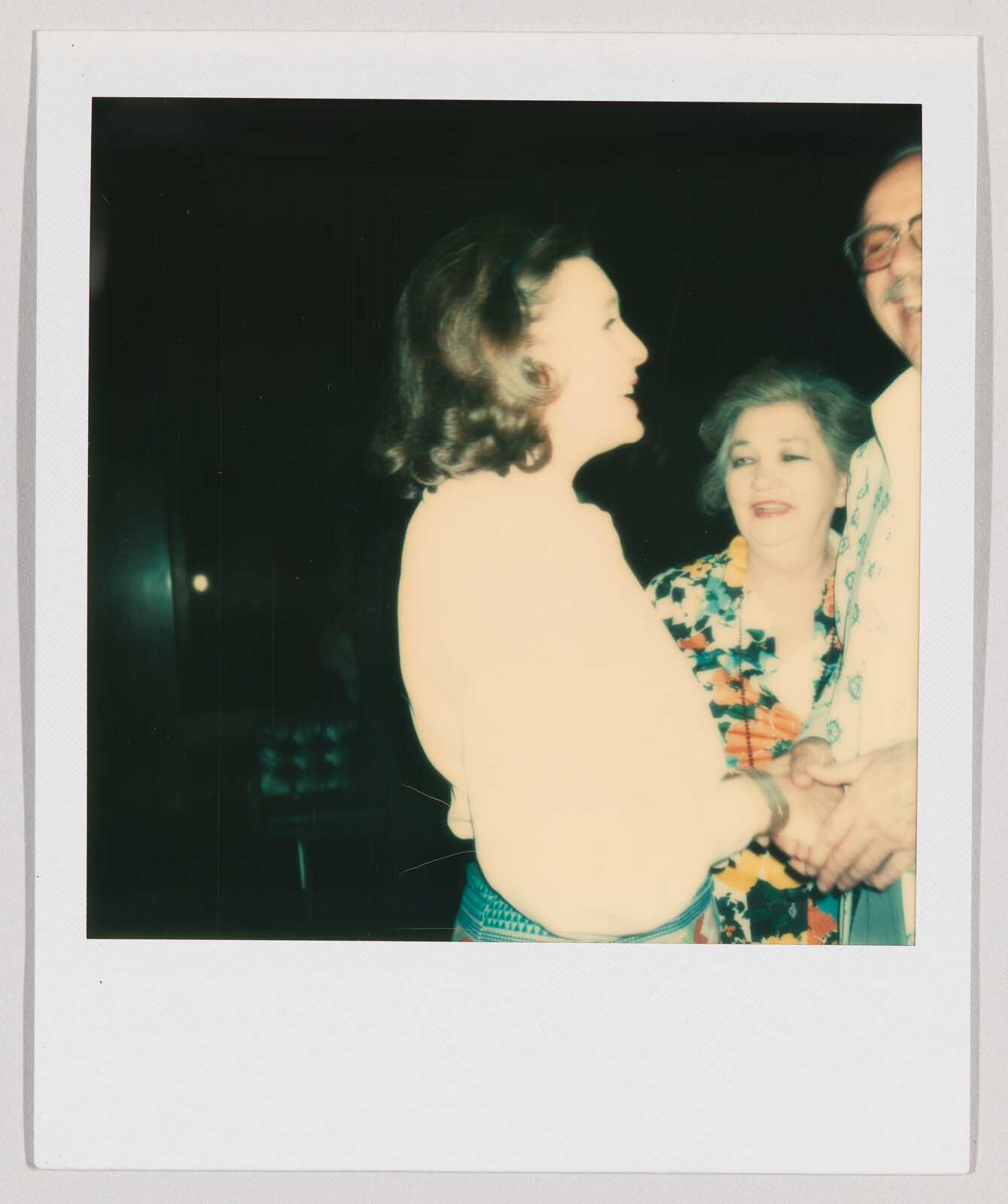 Three adults stand close together, smiling and talking, with one woman holding hands with a man.